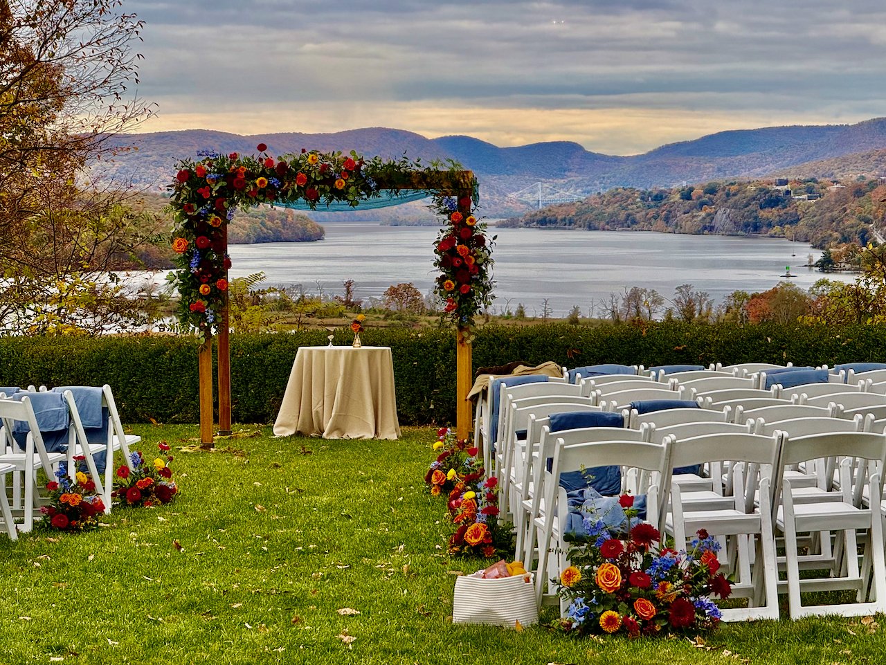 Outdoor wedding setup with white chairs decorated with colorful flowers, a flower-adorned arch over a small table, overlooking a river and mountains in the background.