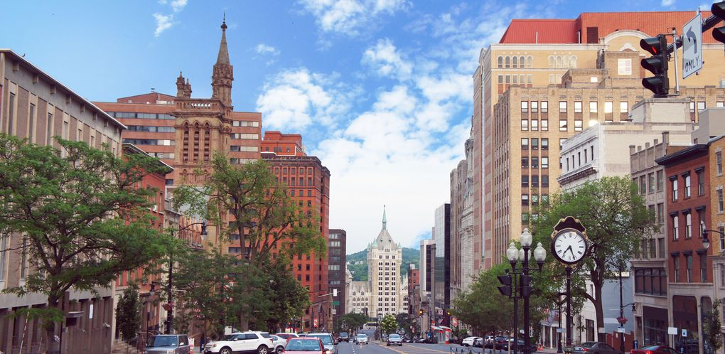 Street view of downtown Salt Lake City with tall buildings, trees, parked cars, a clock, and a blue sky.