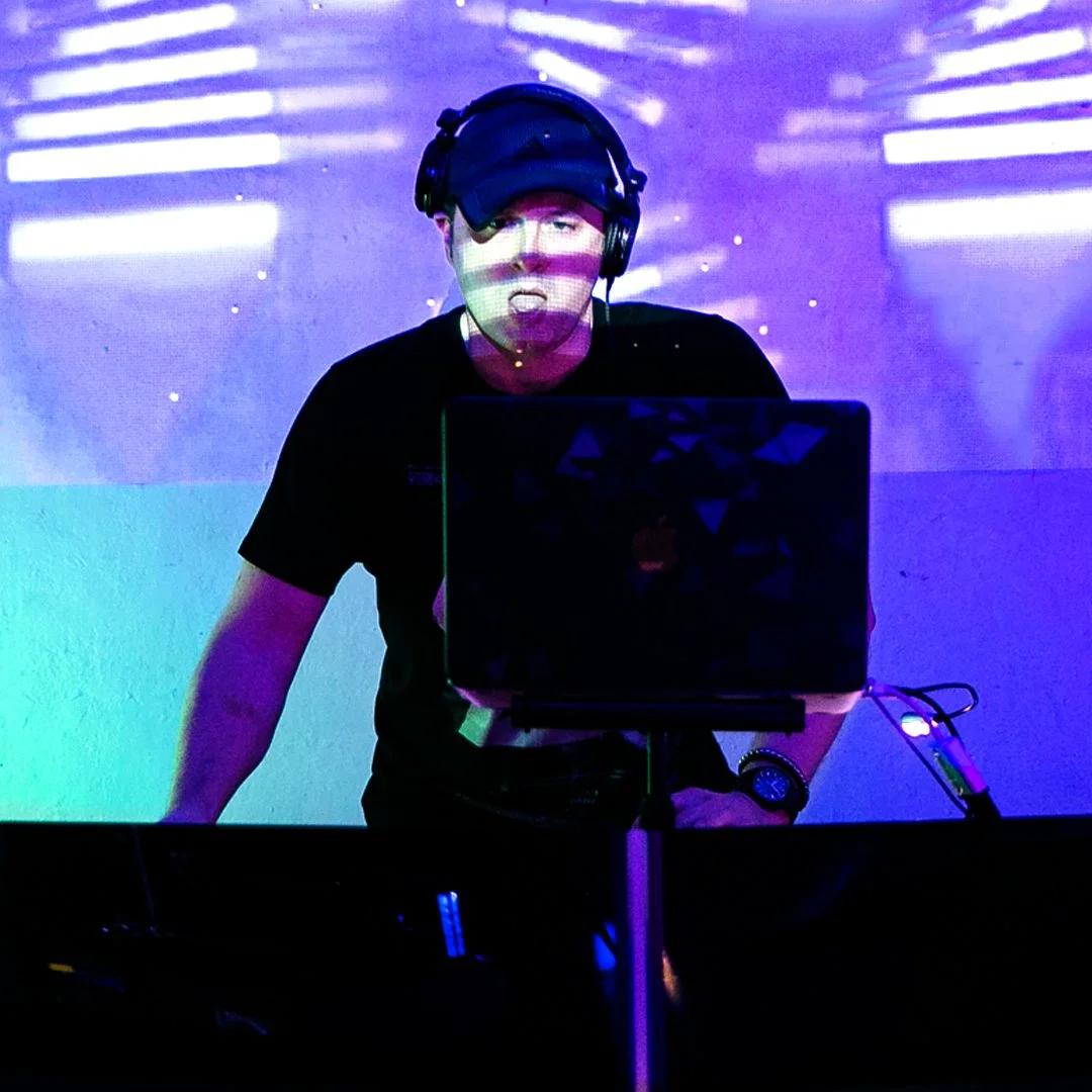 A person wearing a cap and headphones, DJing at a music event with colorful lights and a laptop.