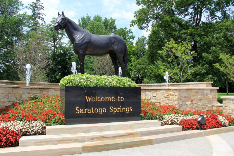 Statue of a horse on a landscaped pedestal with a 'Welcome to Saratoga Springs' sign, surrounded by colorful flowers and water fountains, in a park-like setting with trees in the background.