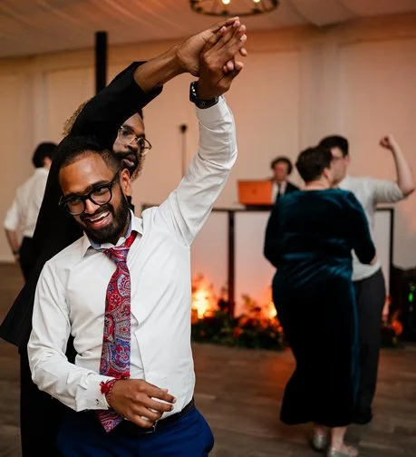 Two men dancing together at a celebration or party, with other people dancing in the background.