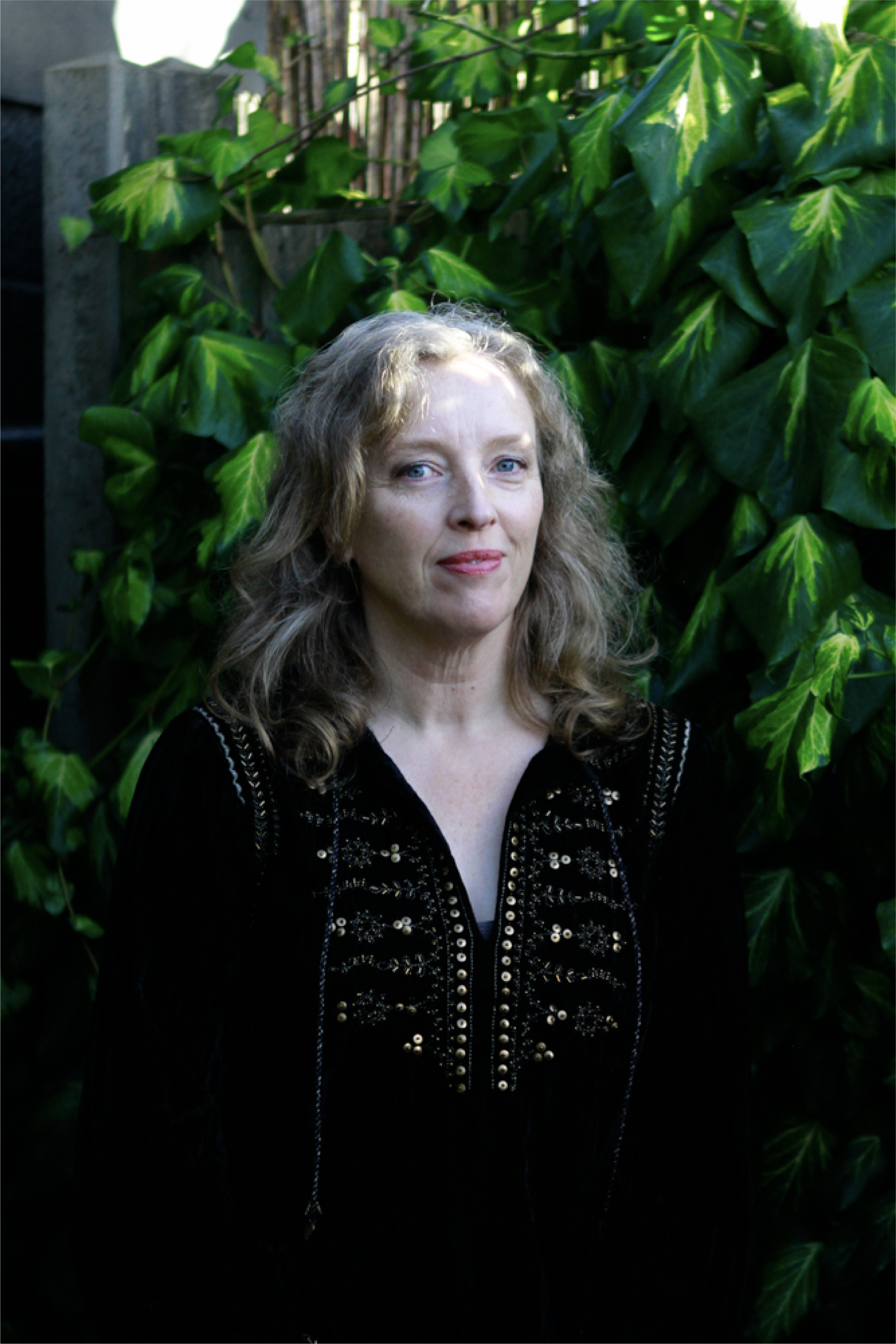 A woman with wavy blonde hair and blue eyes standing outdoors in front of green leafy plants, wearing a black velvet dress with embroidery.