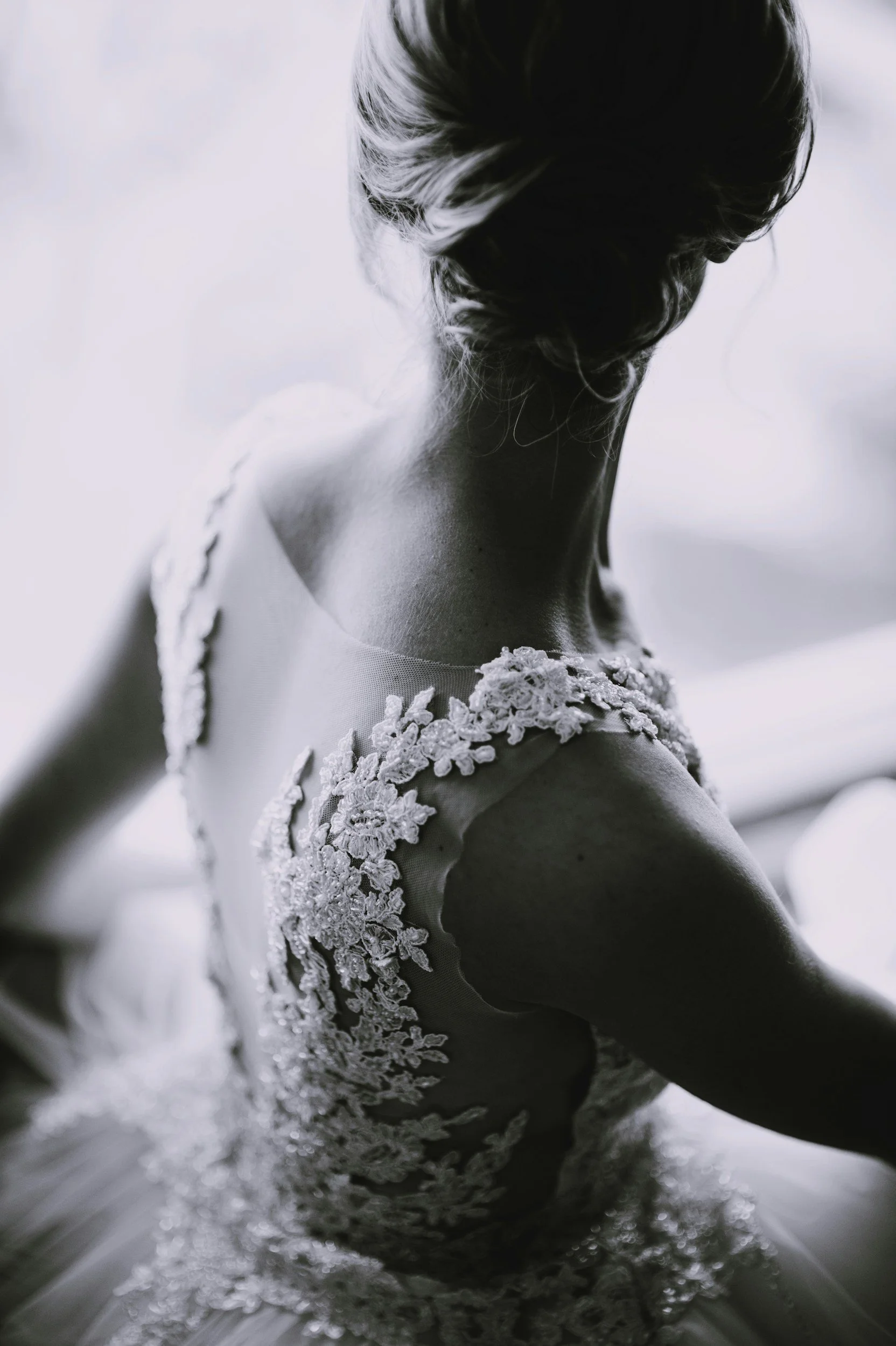 A black and white photo of a woman in a lace dress, viewed from behind with her hair styled in an elegant updo.