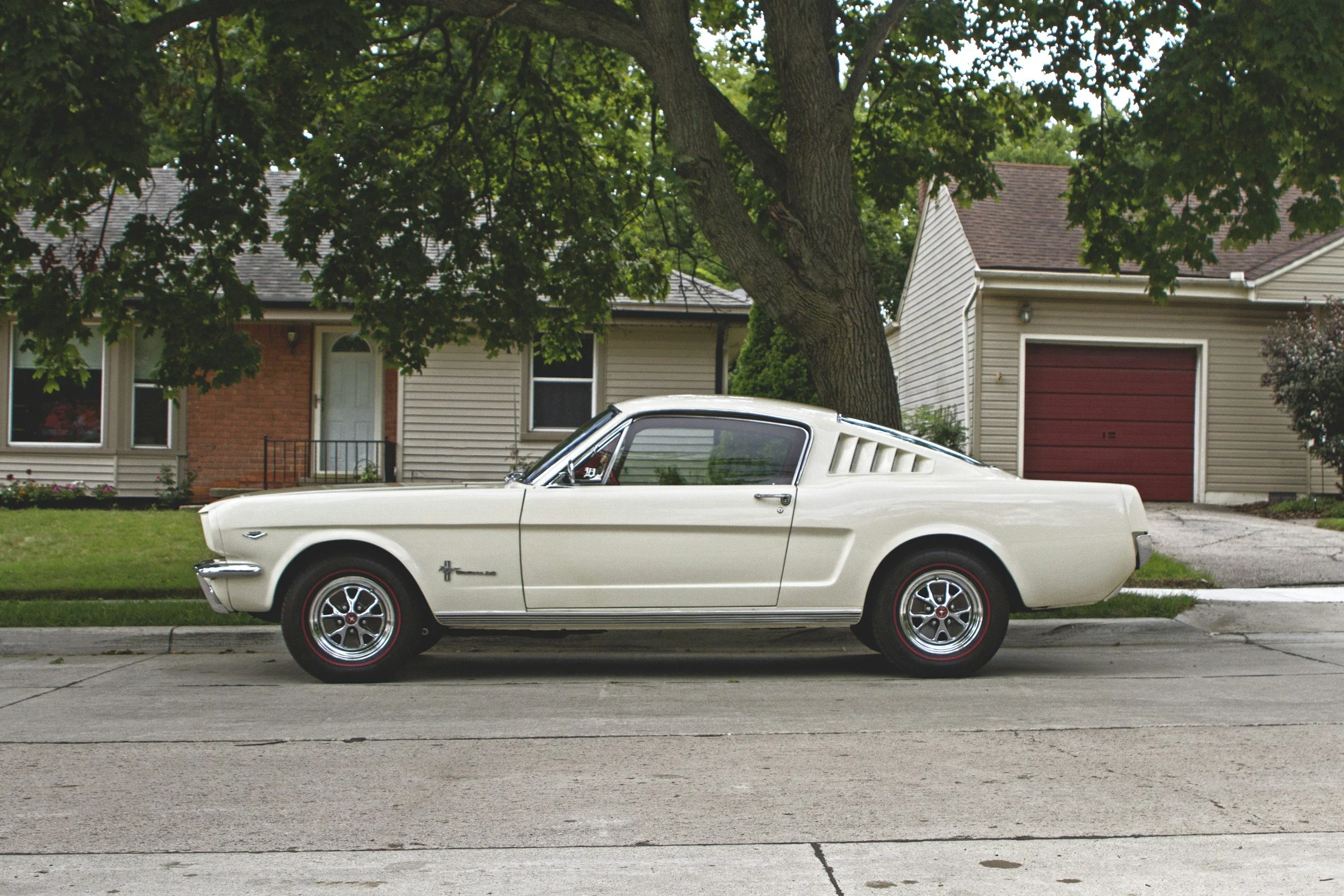 A white vintage Ford Mustang car parked in front of a suburban house with tiled roof and a large tree in the yard.