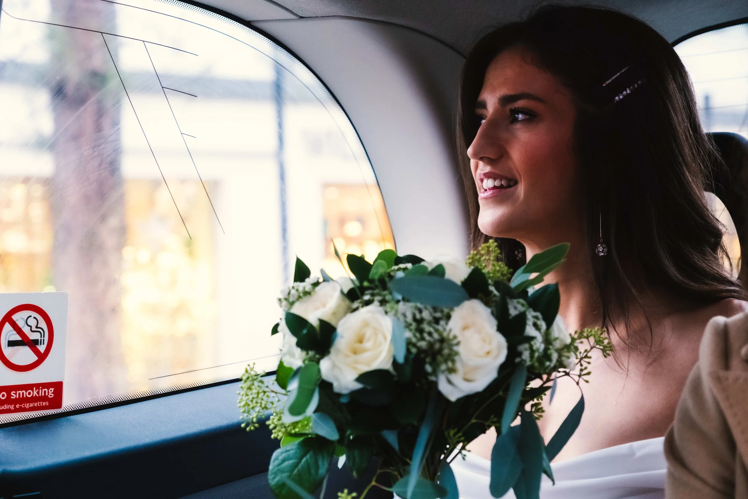 A woman with dark hair and earrings inside a vehicle holding a bouquet of white and green flowers, looking out the window.