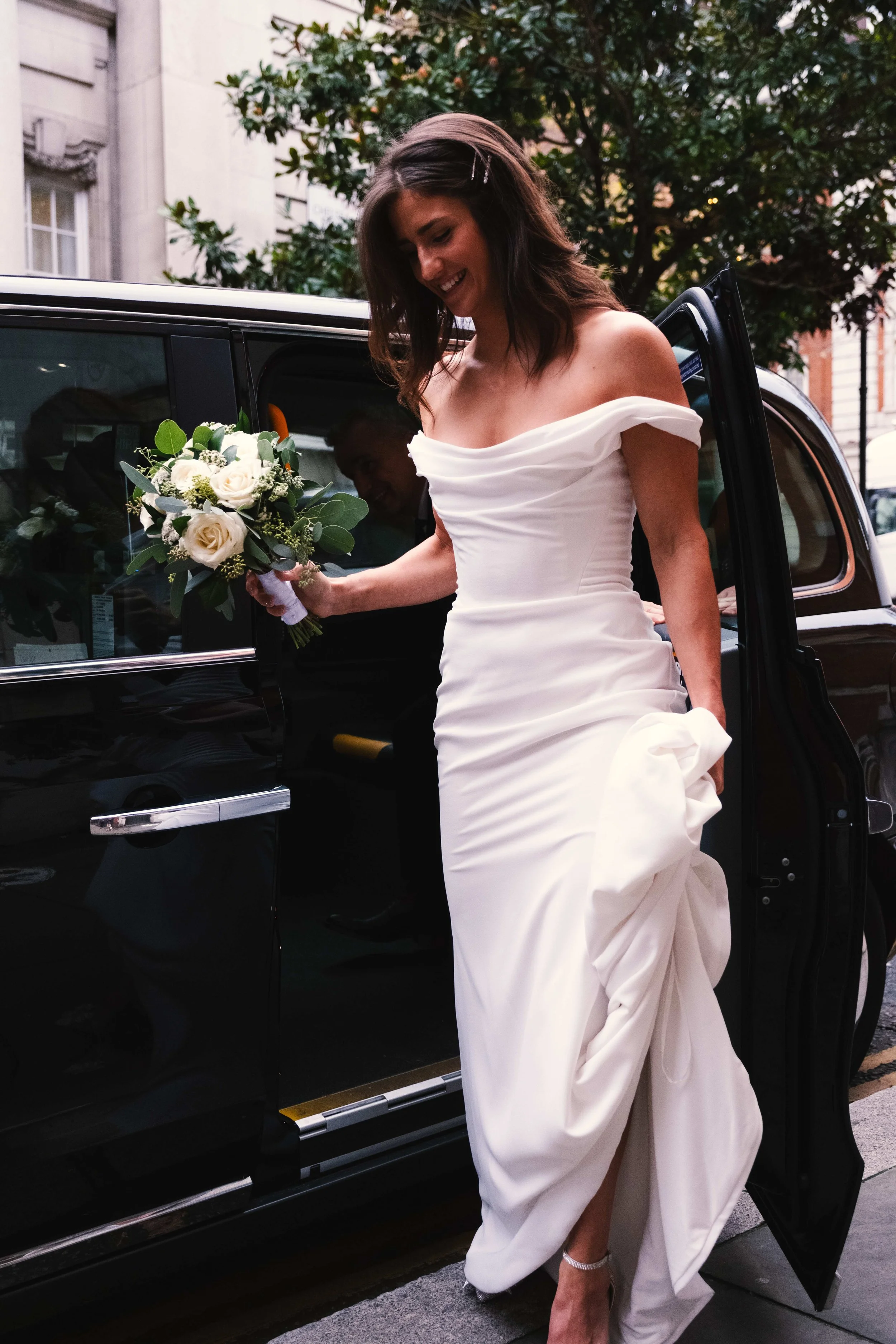 A woman in a white off-the-shoulder gown holding a bouquet of flowers as she exits a black limousine. She is smiling and stepping onto the street, with city buildings and trees in the background.