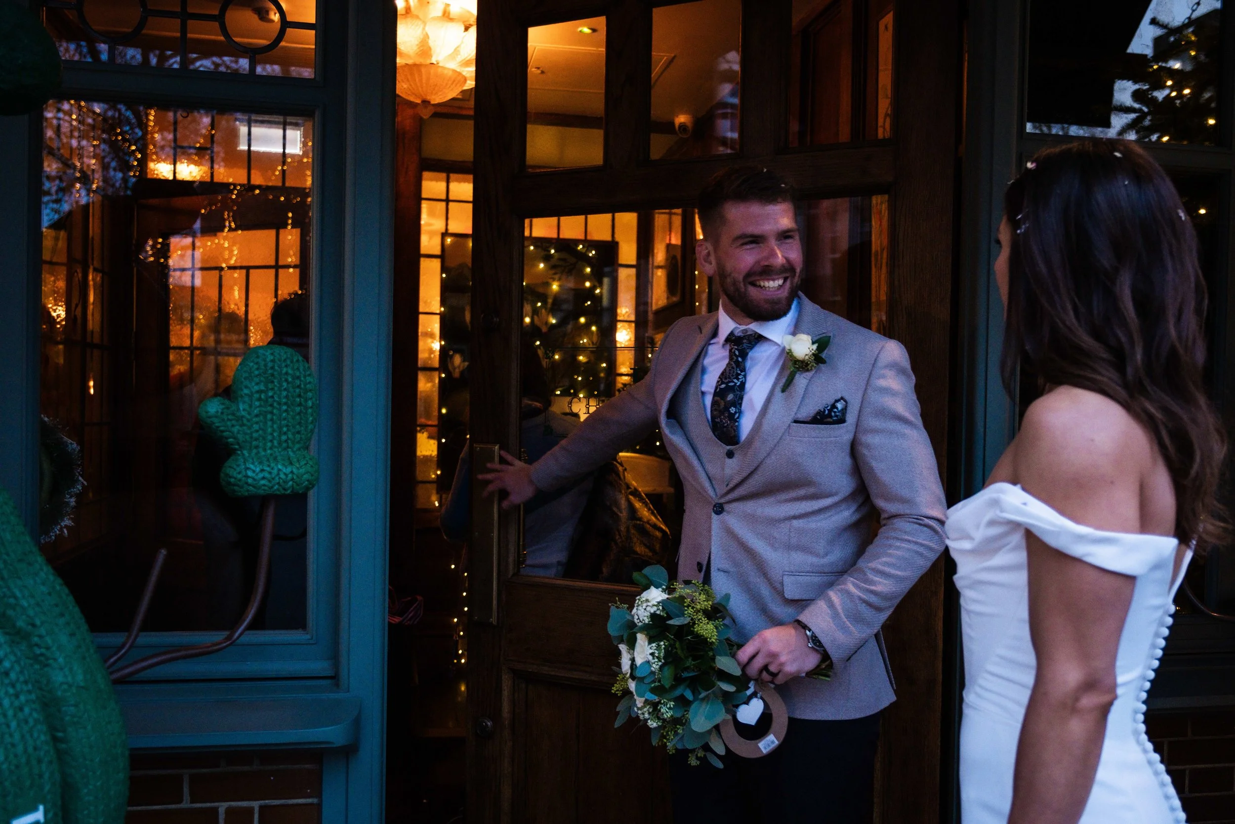 A man wearing a light gray suit with a boutonniere and holding a bouquet of flowers smiling at a woman in an off-shoulder white dress as they converse near a glass door, with warm indoor lighting and festive decorations visible through the window.