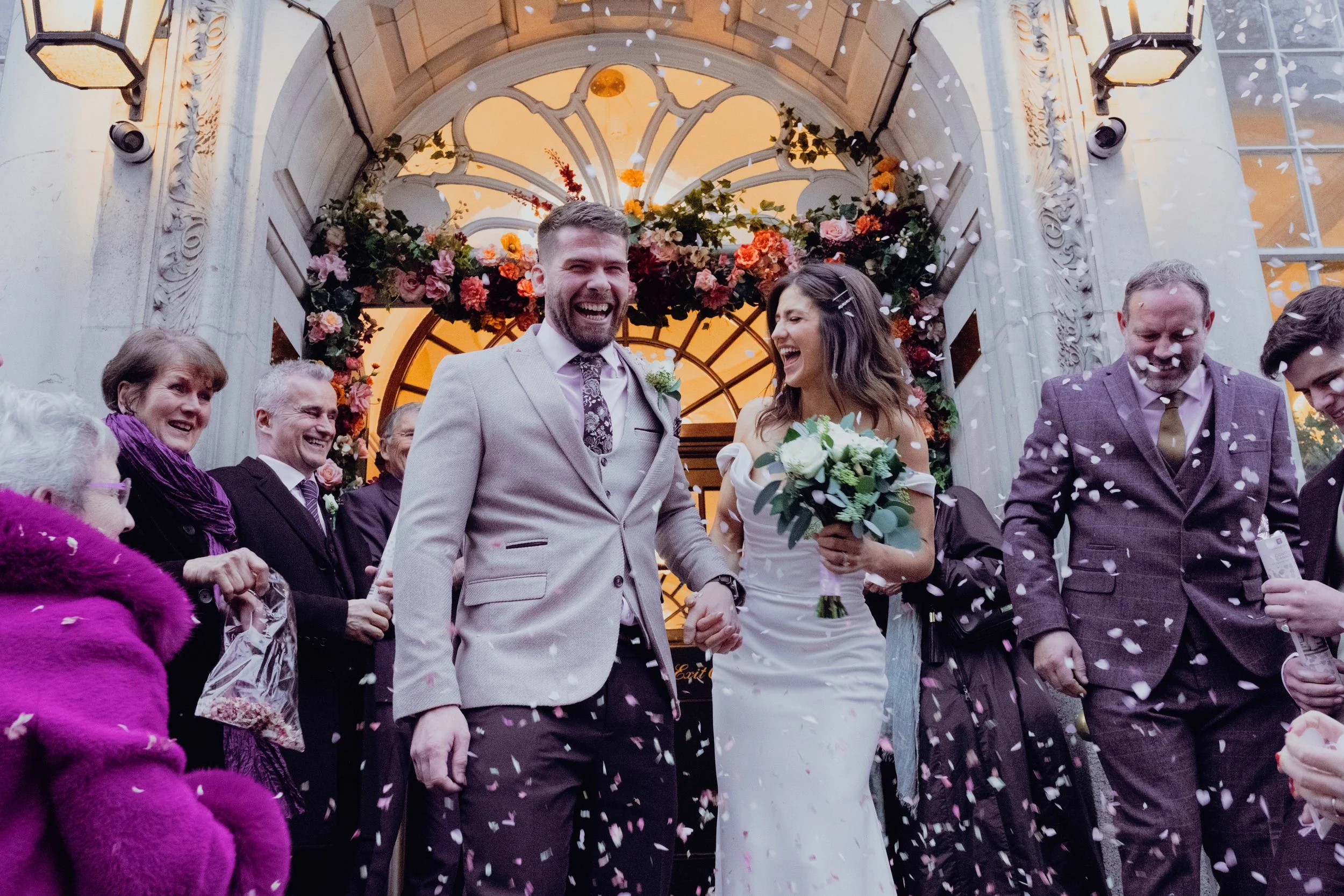 A wedding celebration with a bride and groom holding hands and smiling, surrounded by family and friends indoors, with flowers and confetti.