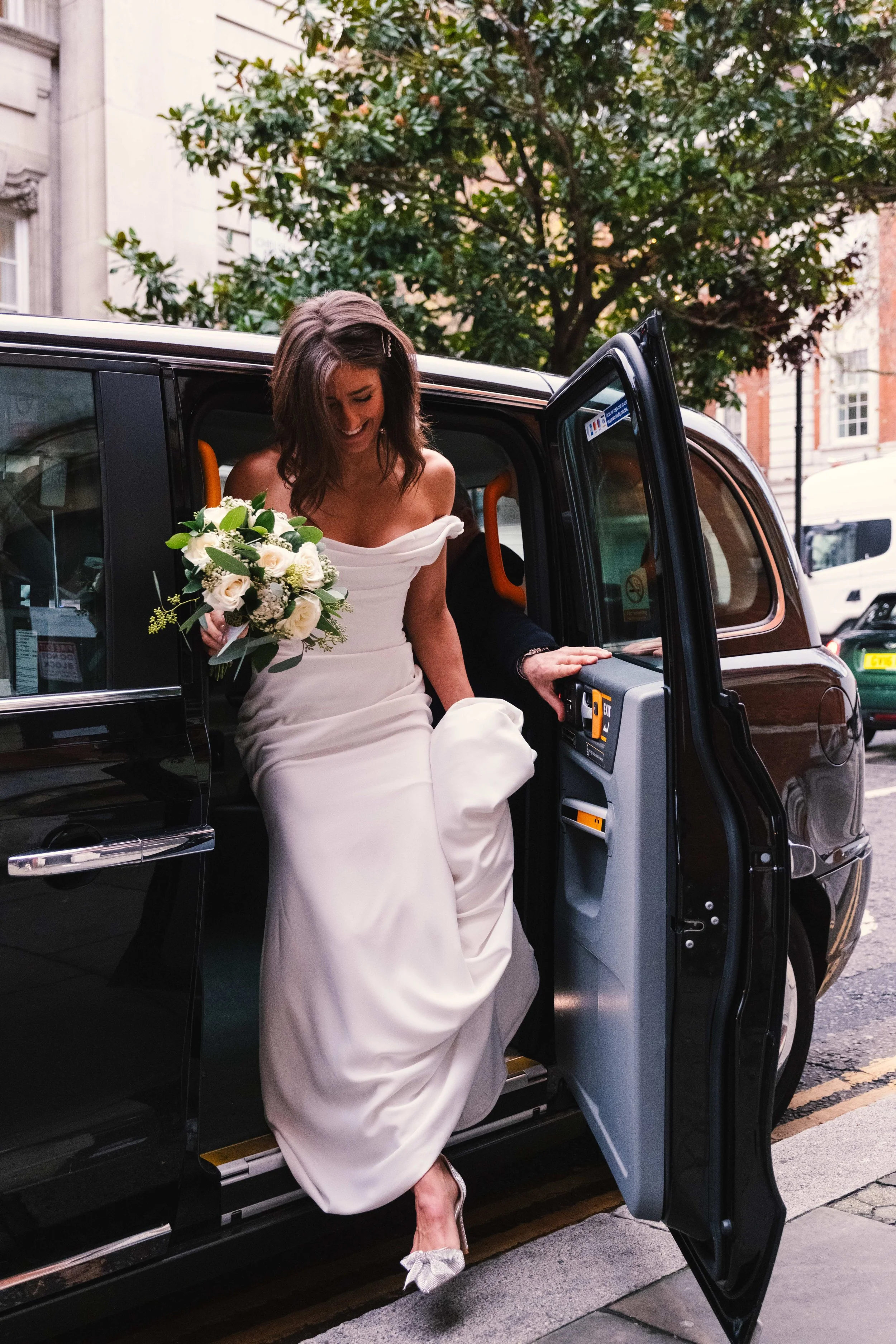 A woman in a white wedding dress holding a bouquet of white roses, stepping out of a black vehicle with a smile on her face.