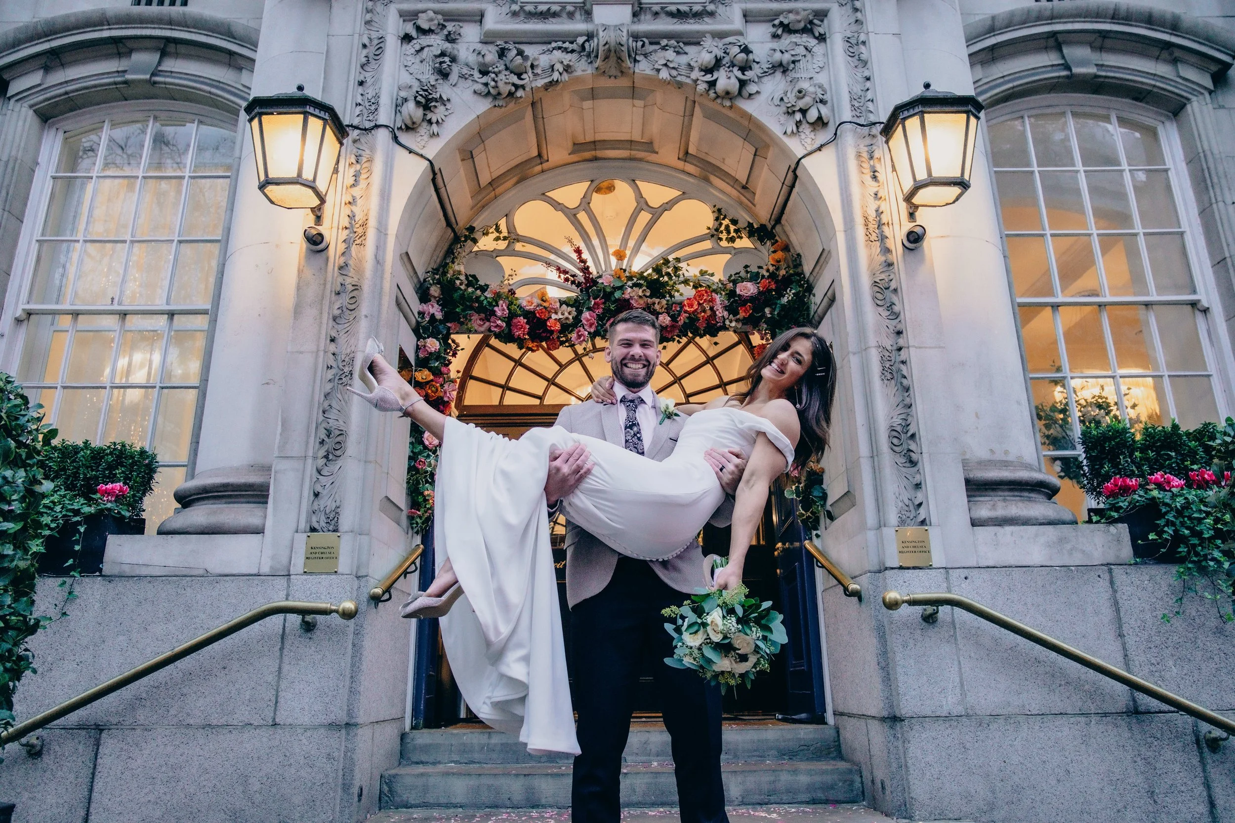 A man in a suit is carrying a woman in a wedding dress down the steps of a building decorated with a floral arch and large windows, outside a historic stone building with ornate carvings and lanterns.