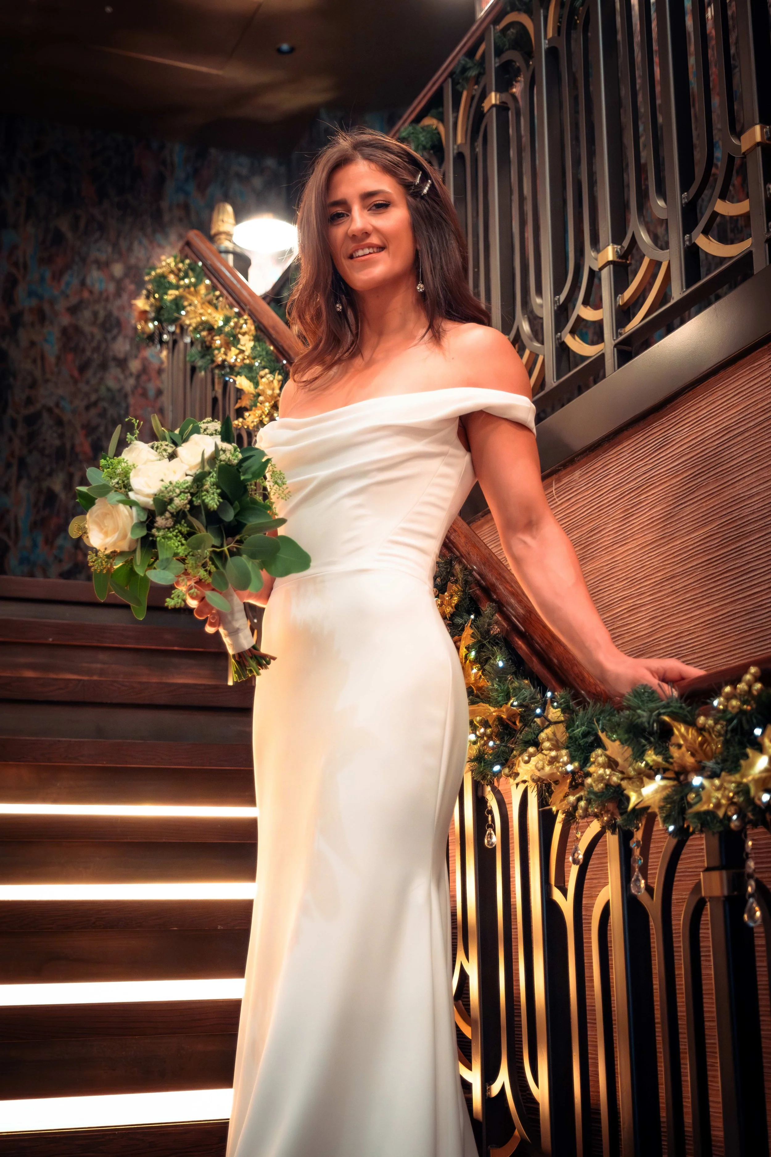 A woman in a white off-shoulder dress holding a bouquet of white roses and greenery, standing on a staircase decorated with Christmas garland and lights, with a decorative metal railing behind her.