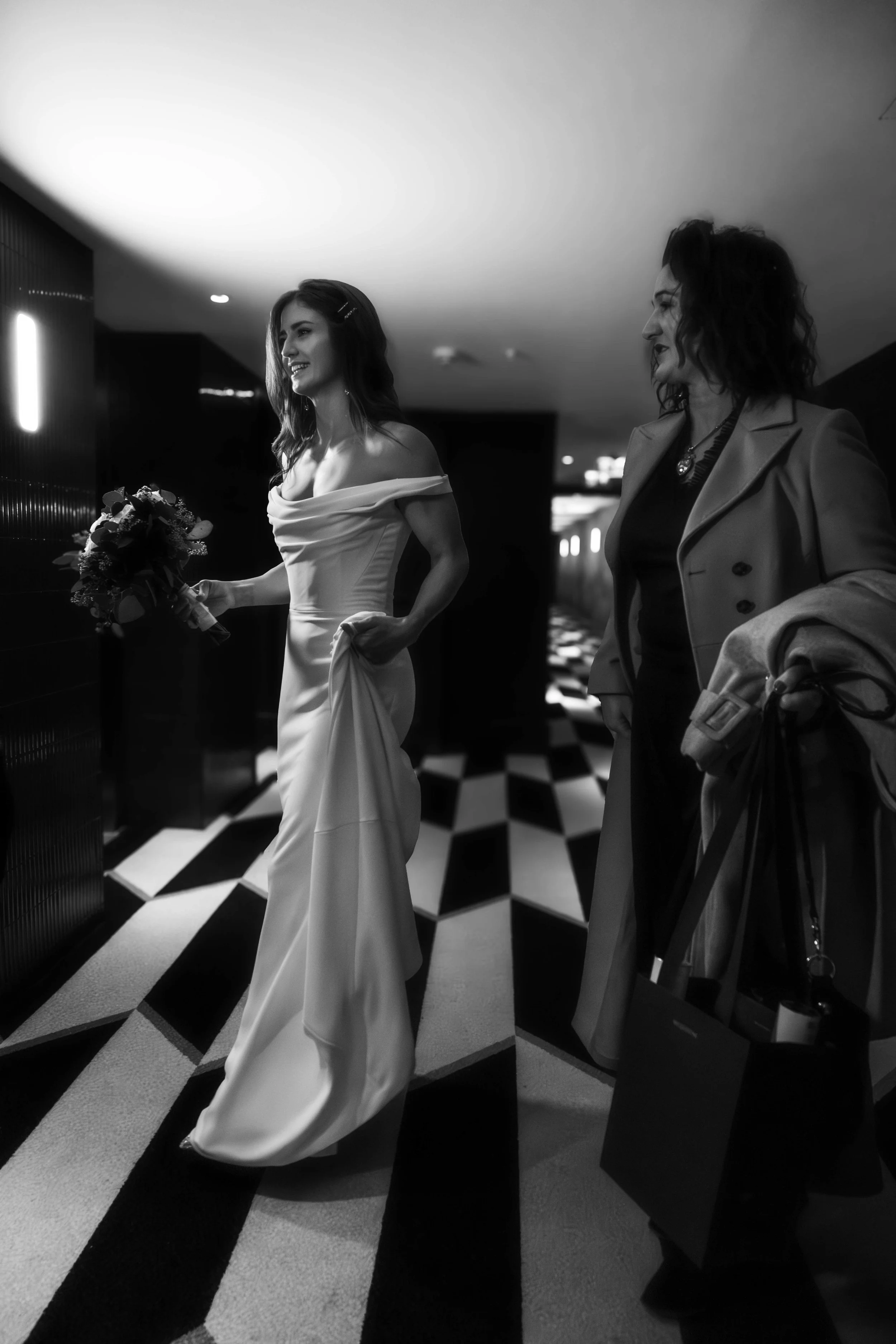 A woman in a white off-shoulder dress holding a bouquet of flowers walking in a modern hallway, smiling, with another woman in a blazer standing nearby, smiling.