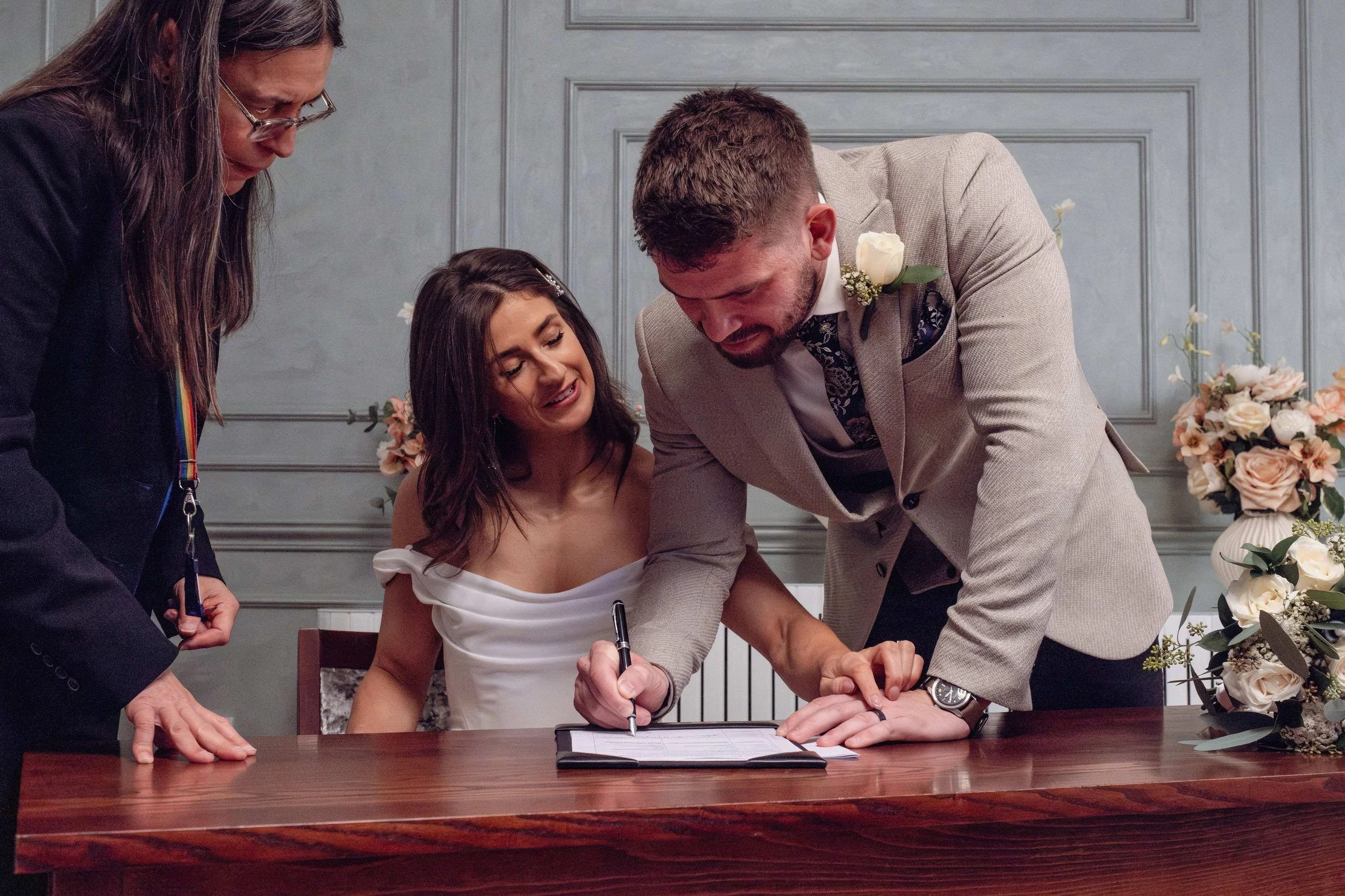 A woman in a white dress signs a document at a wedding ceremony, surrounded by people, with floral arrangements in the background.