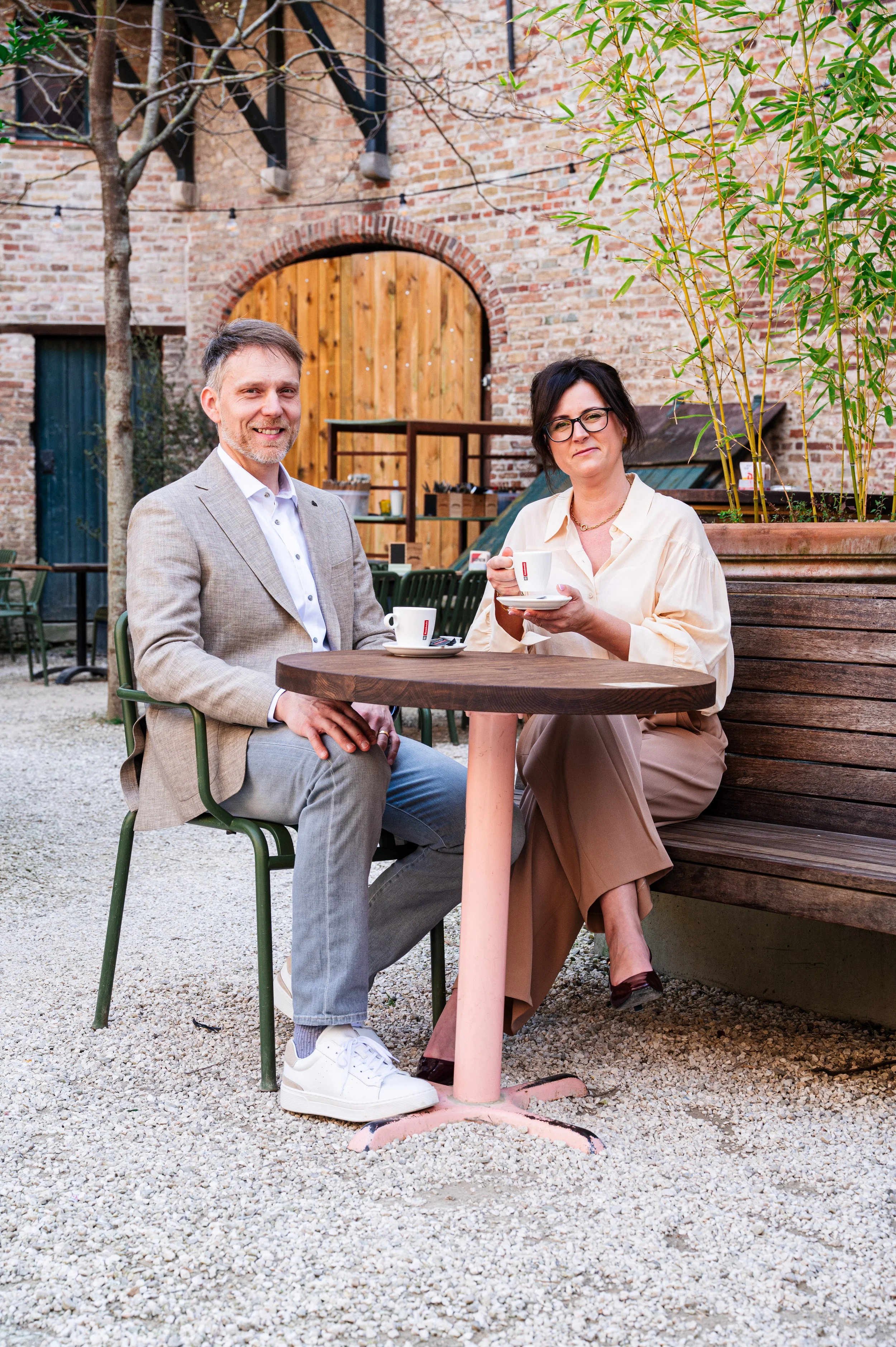 Twee mensen, Kurt en Stefanie, zitten aan een buiten tafel van een café of restaurant, een man in een lichtgrijze zakelijke jas en een vrouw in een beige blouse en brede broek. Ze drinken koffie. Ze zitten op een terras omgeven door planten en bomen.