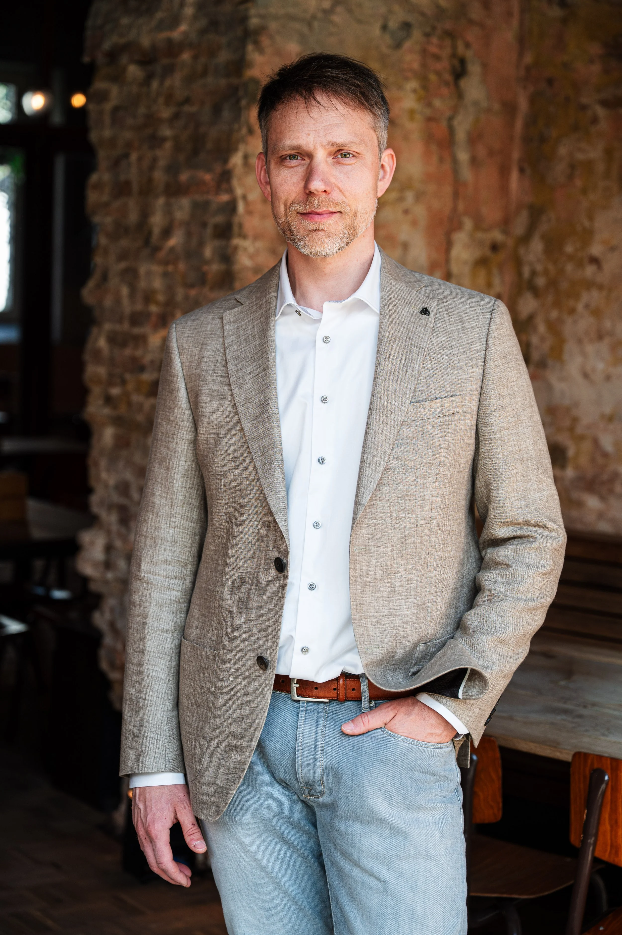 Man, Kurt Braem van Coerant, in een grijs pak met witte blouse en blauwe jeans, staand in een interieur met stenen muur, korte haren en een lichte baard, met één hand in de broekzak.