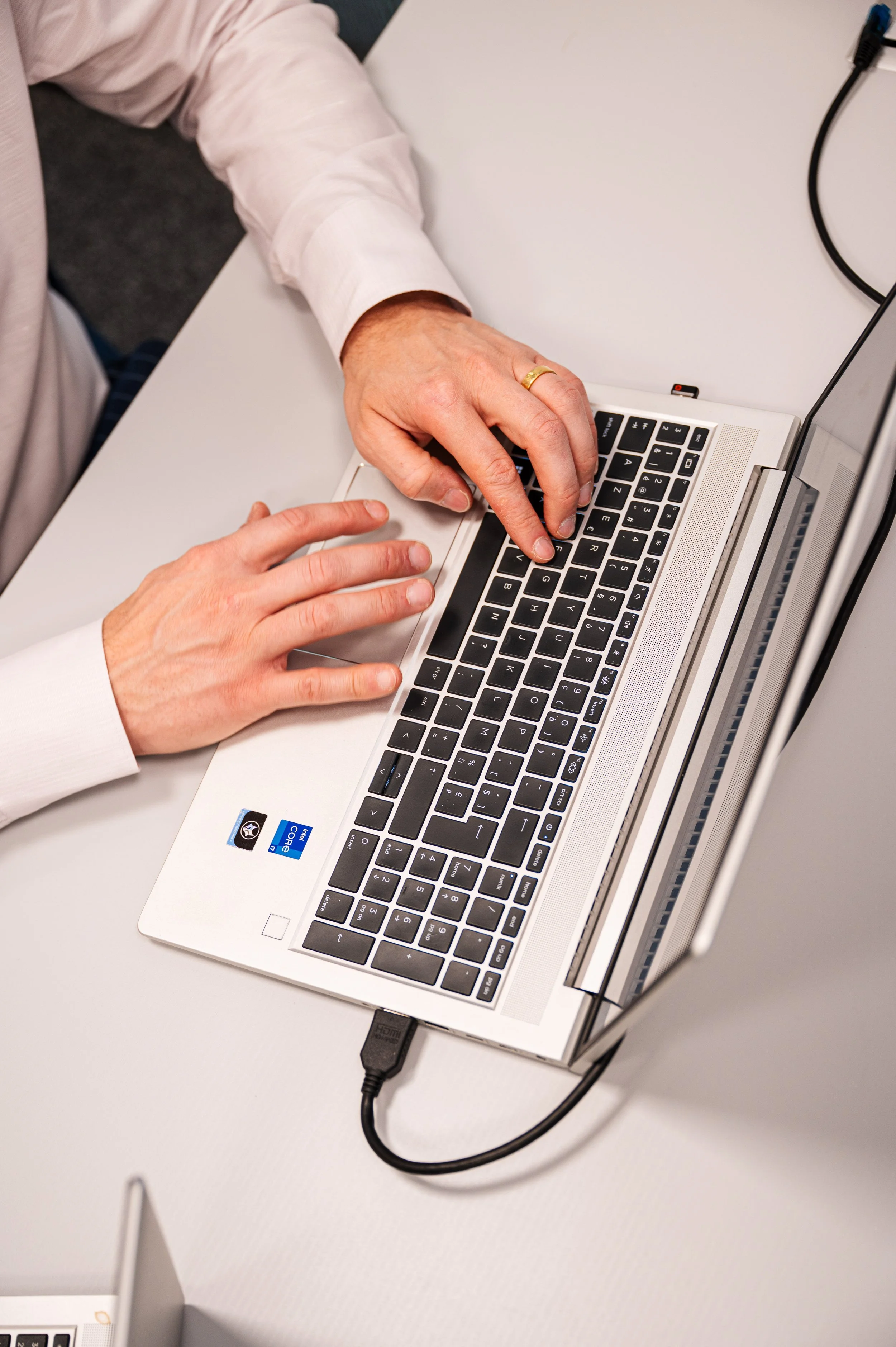 Een close-up van een hand die op een laptoptoetsenbord typt, met een andere hand die de muis bedient. De laptop ligt op een wit bureau.