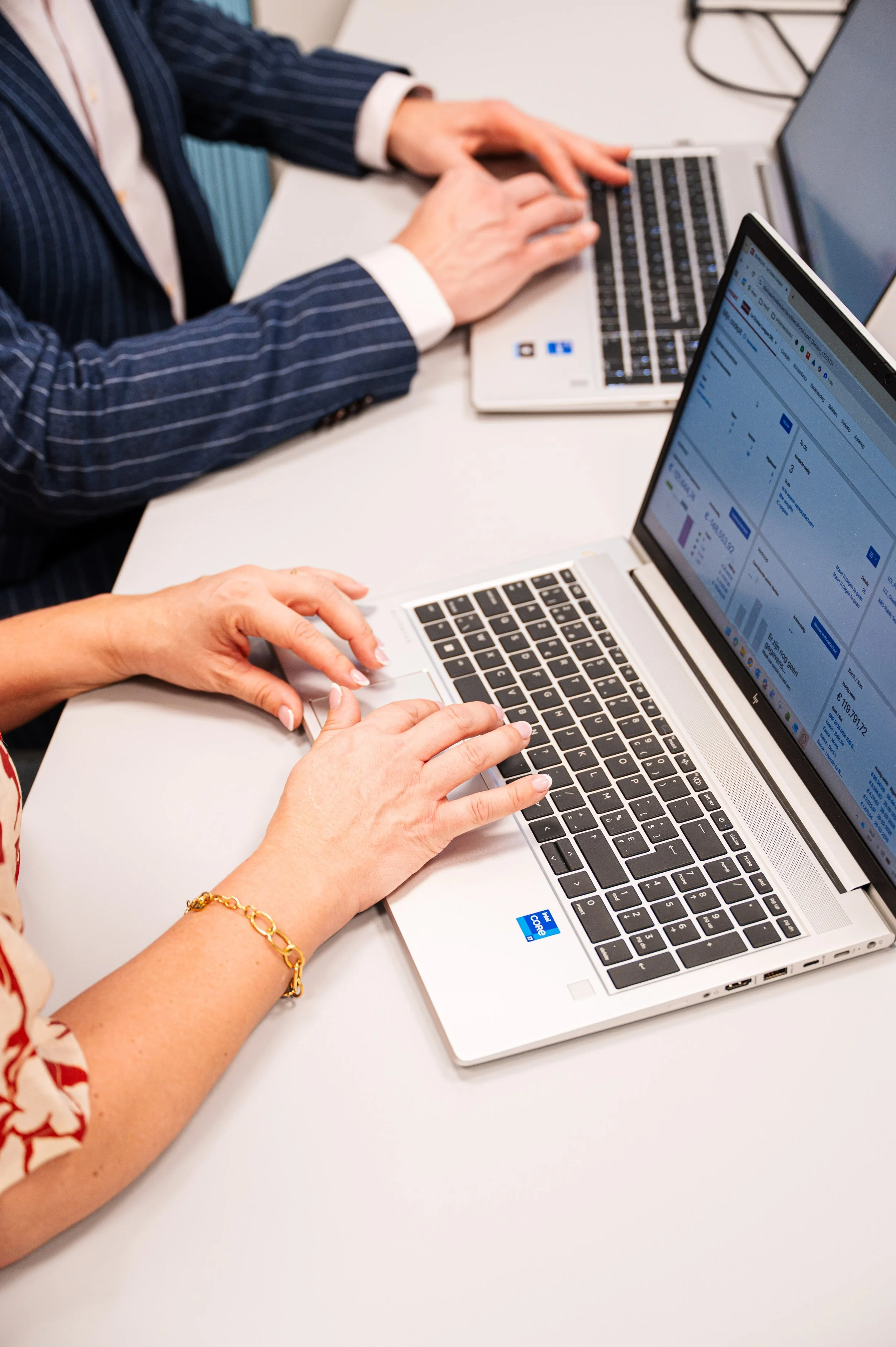 Twee mensen, Kurt en Stefanie van accountantskantoor Coerant, werken aan laptops op een witte tafel, één persoon met een gouden armband en een persoon met een blauwe gestreepte jas.