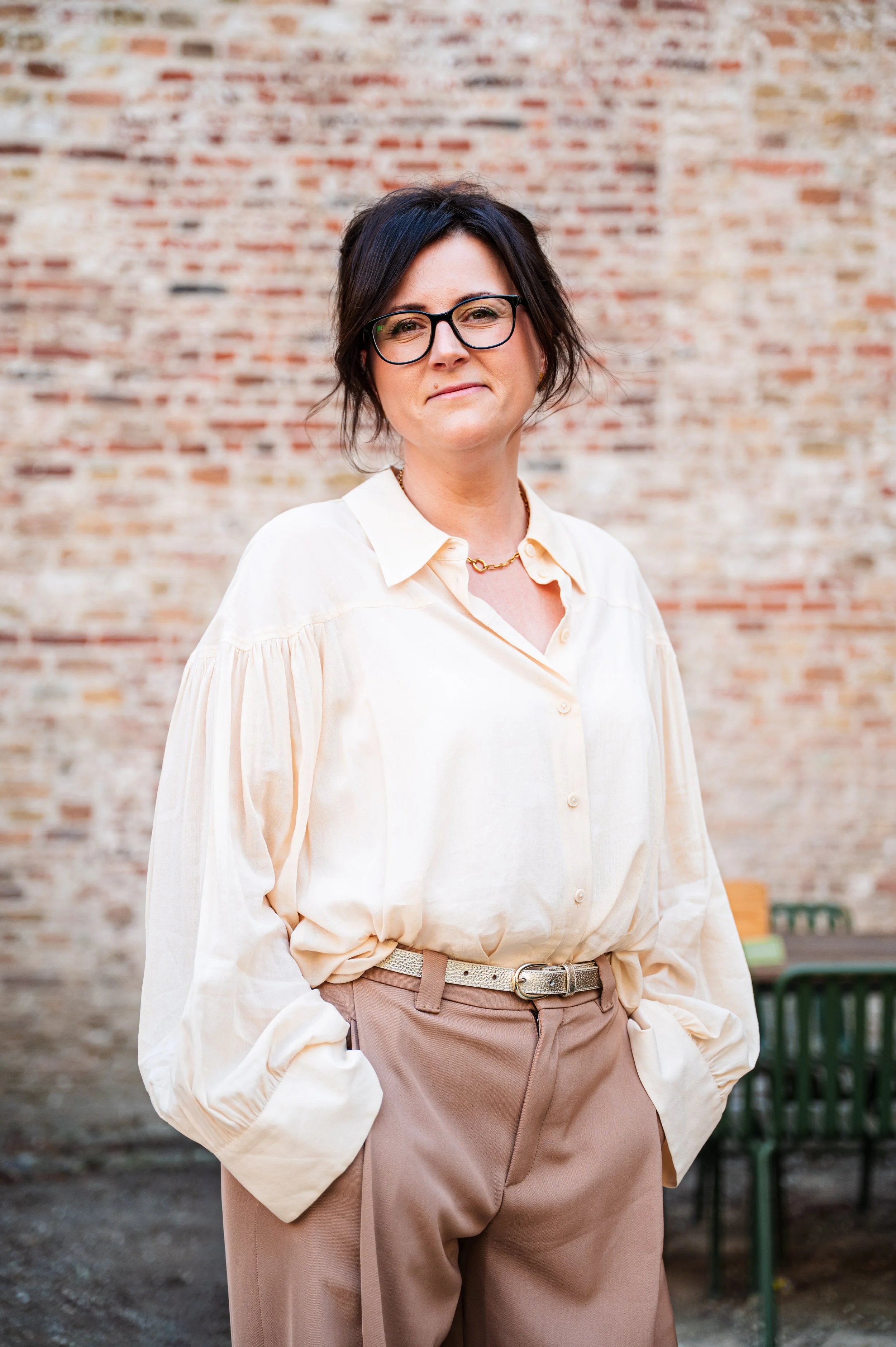 Vrouw, Stefanie Lareu van Coerant, met korte, donkere haar en bril, gekleed in een lichte blouse en beige broek, staande buiten voor een bakstenen muur.