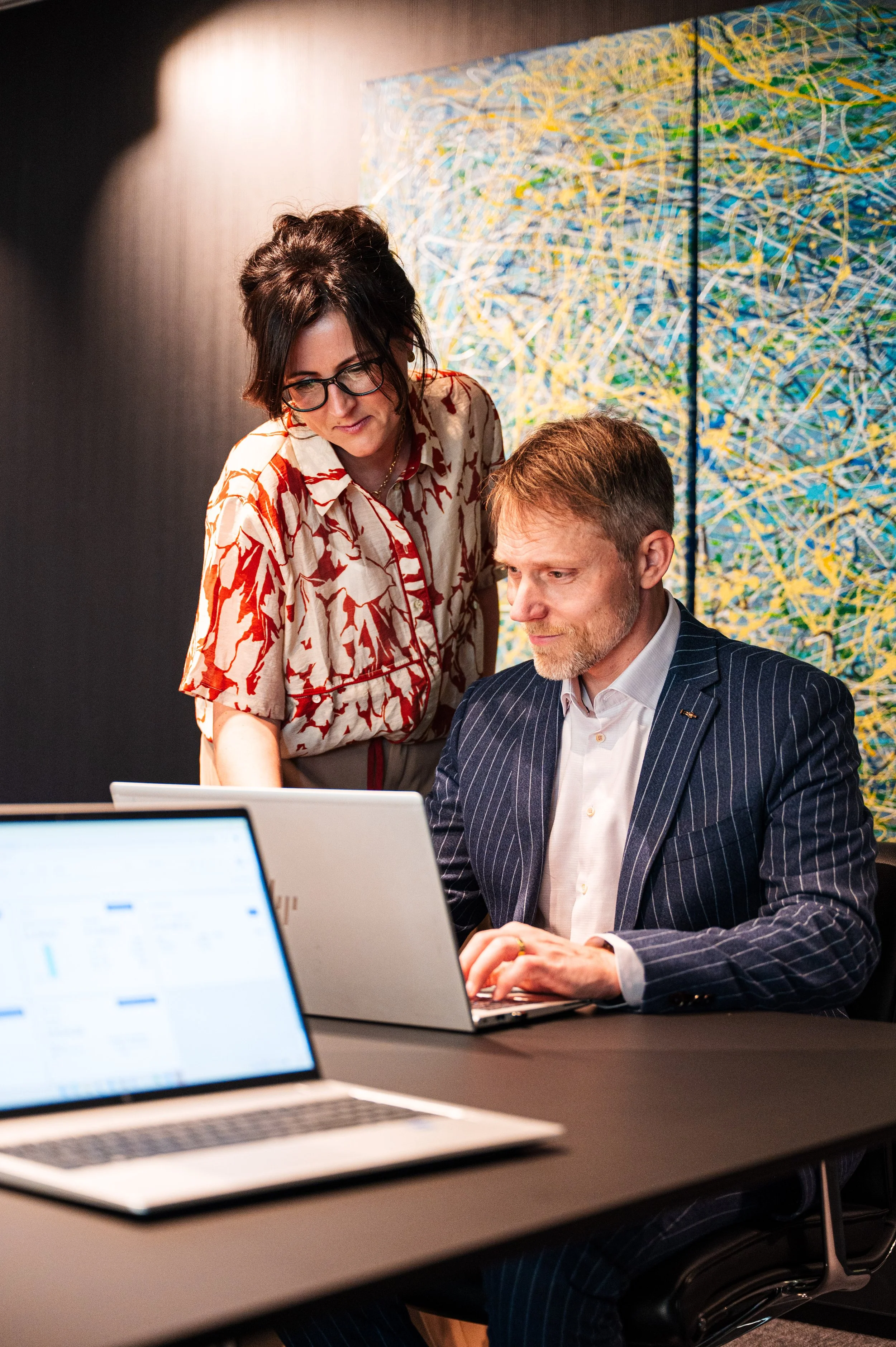 Een vrouw, Stefanie, met bruin haar, in een blouse met een rood-wit patroon, staat naast een man, Kurt, met een grijze baard en haren, gekleed in een donkerblauw, gestreept pak, die op een laptop werkt. Ze lijken samen te overleggen of te bespreken.