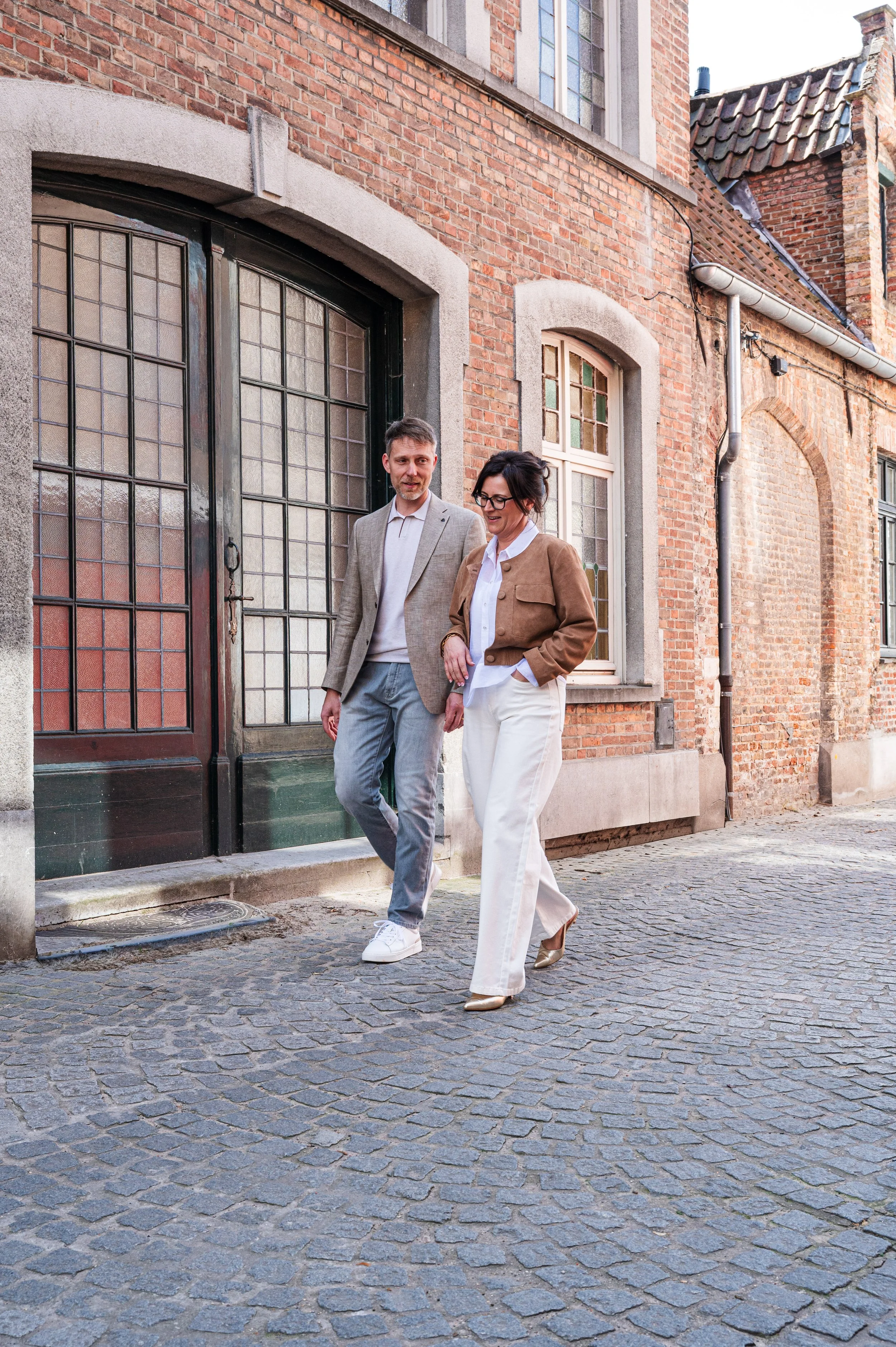 Twee mensen, Kurt en Stefanie van accountantskantoor Coerant, lopen op een oude stenen straat in een stad met bakstenen gebouwen en grote ramen. Ze lijken te genieten en hebben vriendelijke uitdrukkingen.