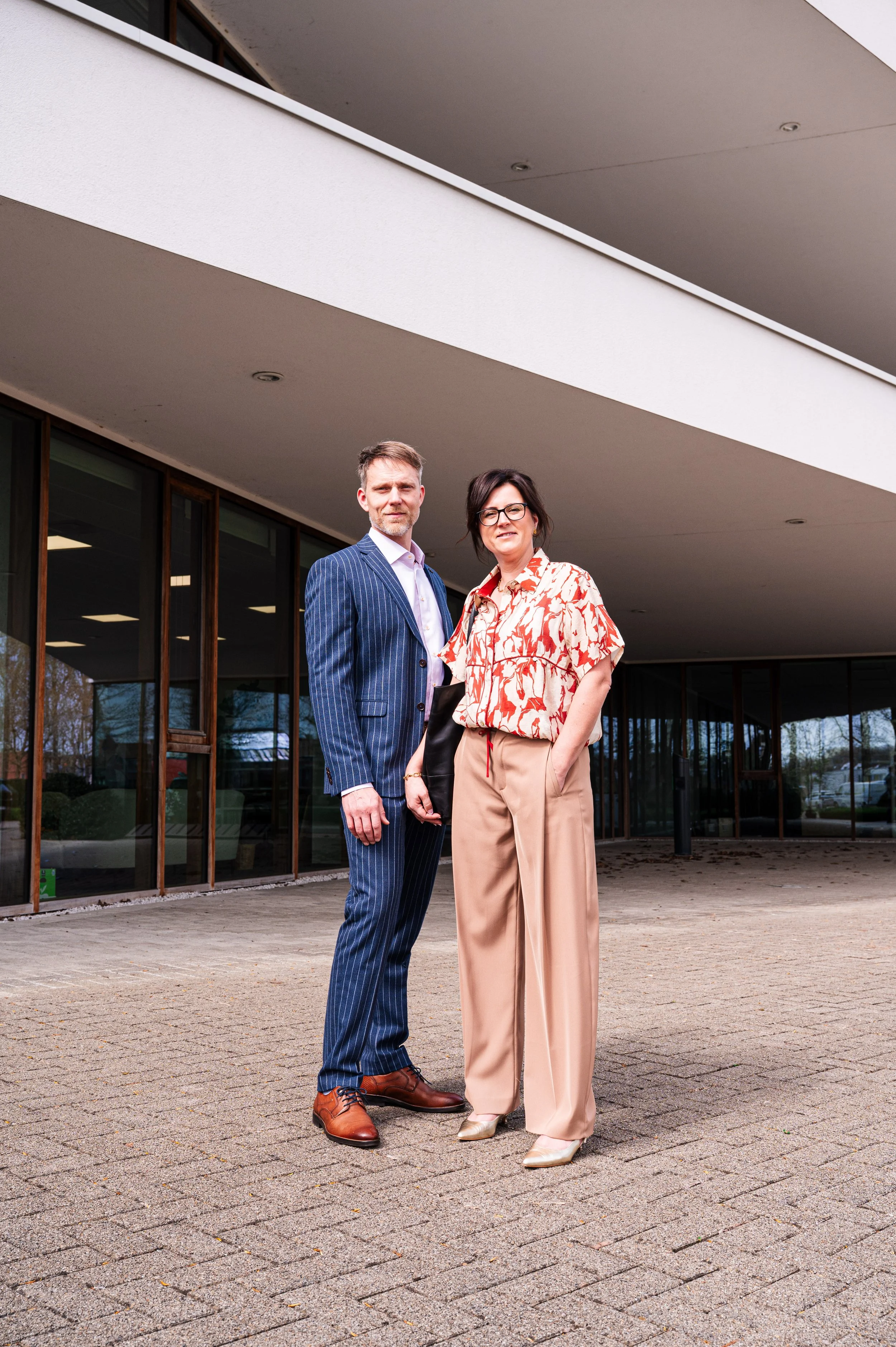 Twee zakenmensen, Kurt en Stefanie van accountantskantoor Coerant,  staan buiten voor een modern gebouw, de man draagt een blauw gestreept pak en de vrouw draagt een beige broek en een blouse met een bril, ze kijken naar de camera.