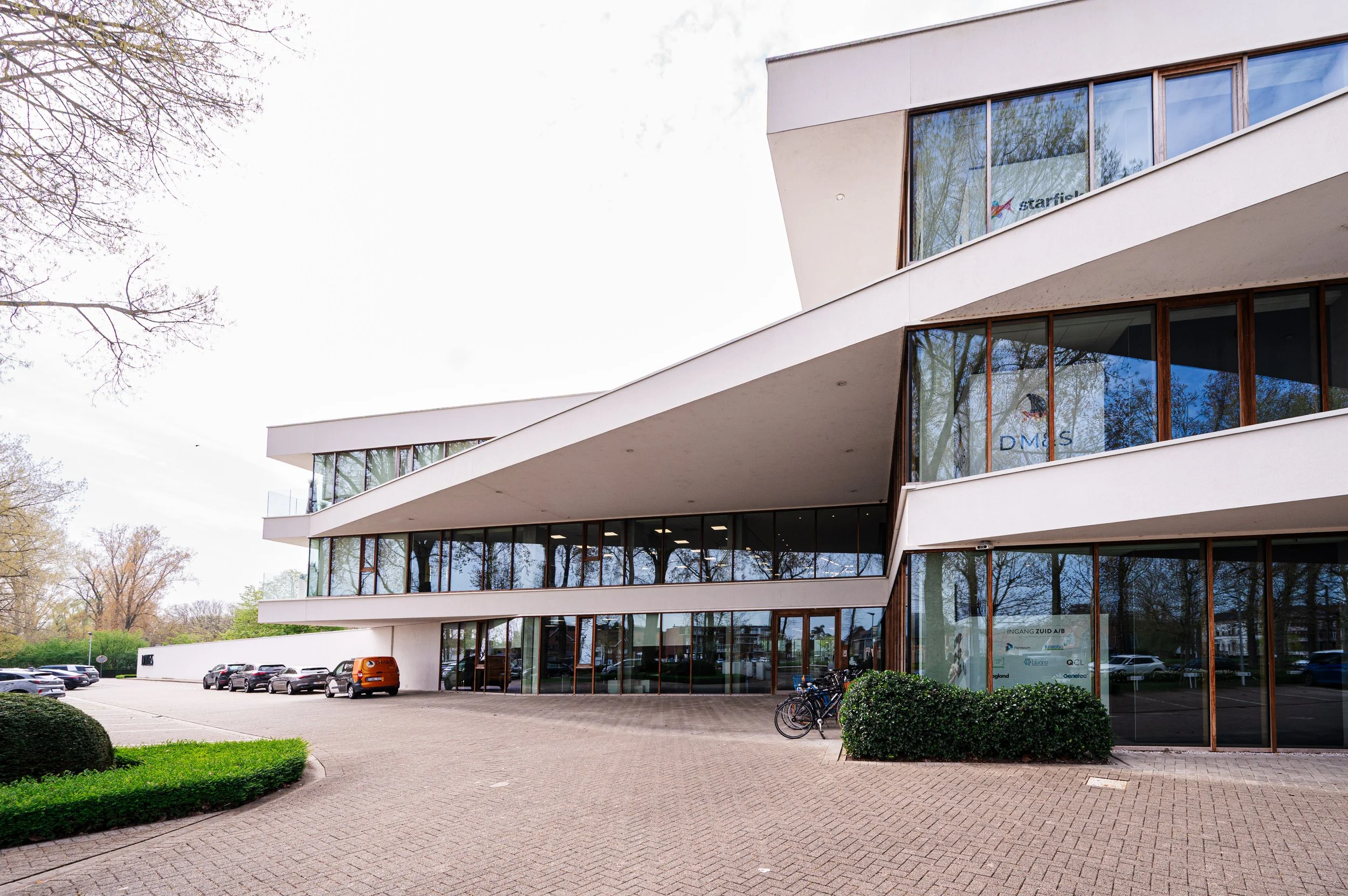 Een modern wit gebouw met grote glasramen en een parkeerterrein met auto's voor. Bomen en een fietsenstalling zijn zichtbaar rondom het gebouw.