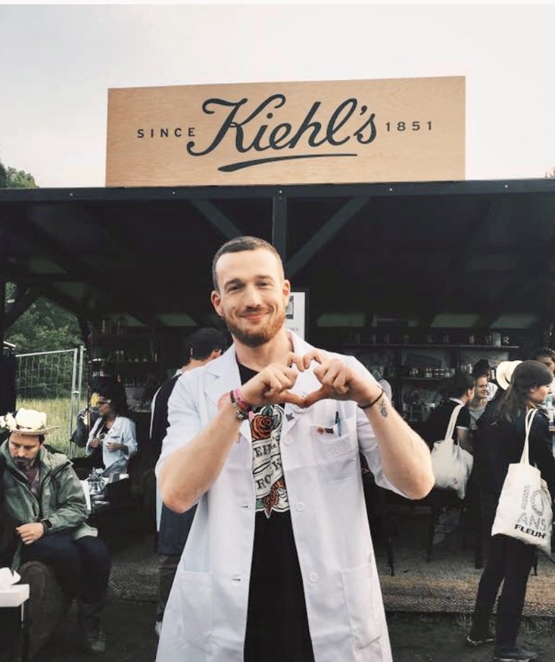 Mathias Landier souriant en blouse blanche faisant un cœur avec ses mains lors d'un événement en plein air, devant un stand Kiehl's.