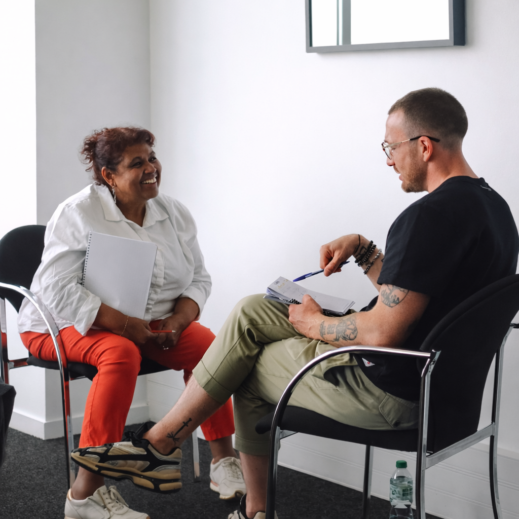 Une femme et un homme discutent dans un bureau, assis sur des chaises noires, près d’un mur blanc avec une fenêtre visible en haut.