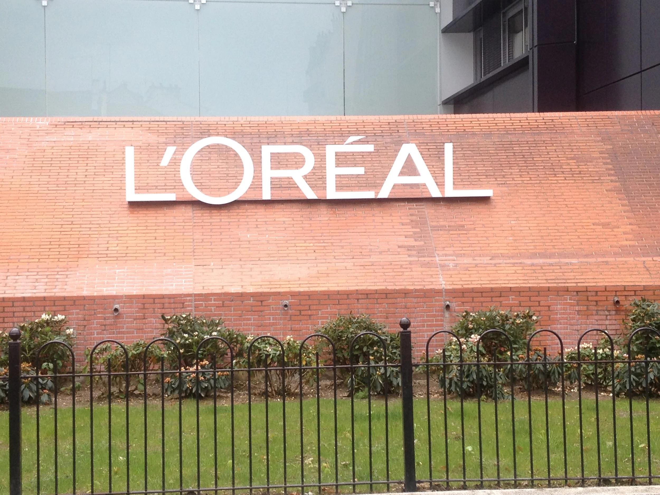 Mur en briques rouges avec le logo blanc de L'Oréal, une petite pelouse verte en premier plan, une barrière noire et des buissons avec des fleurs.