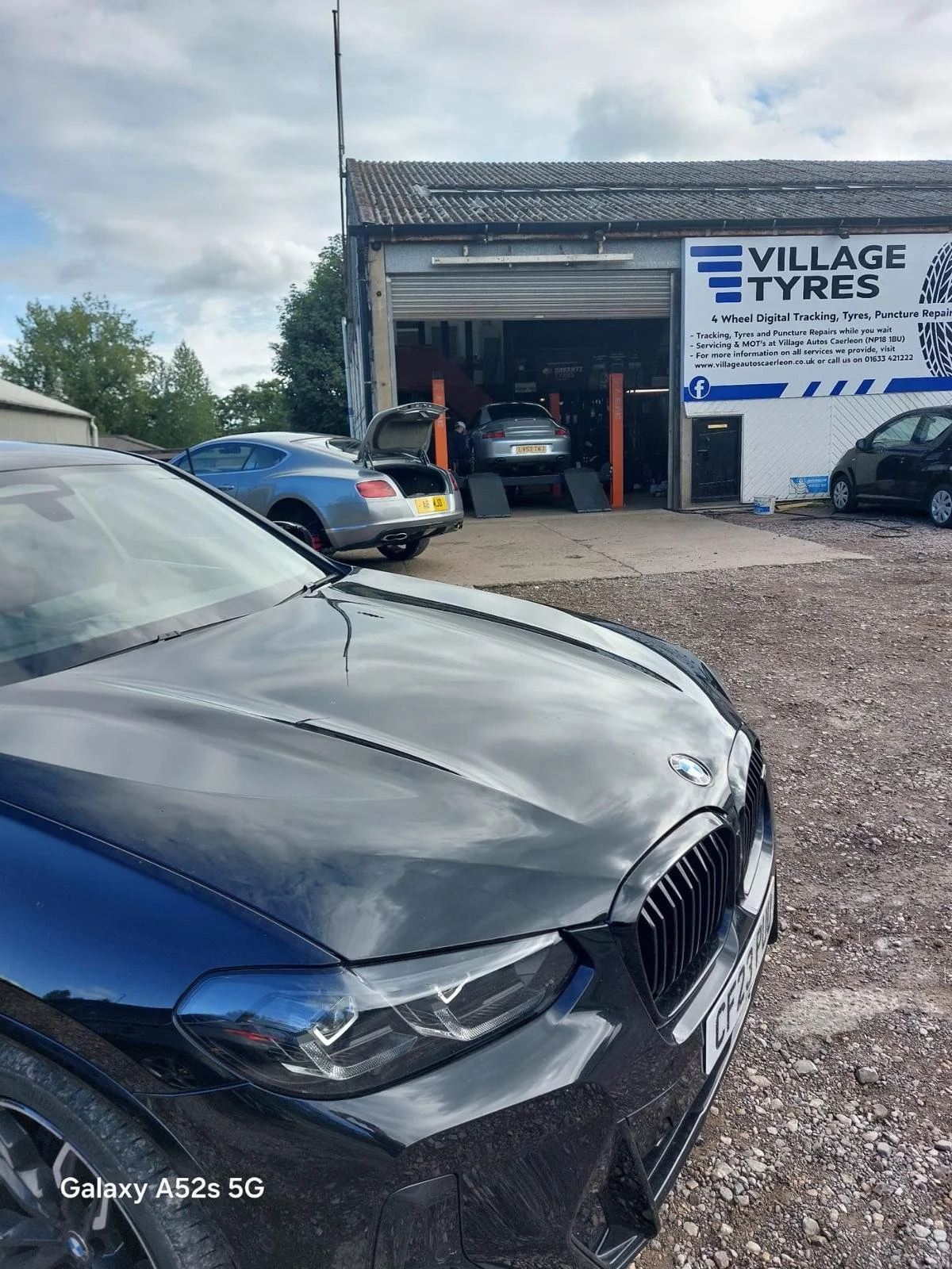 Several cars parked outside a tire shop with a car lifted on a hydraulic lift inside. The shop has a white and blue sign that reads 'Village Tyres.' The sky is partly cloudy.