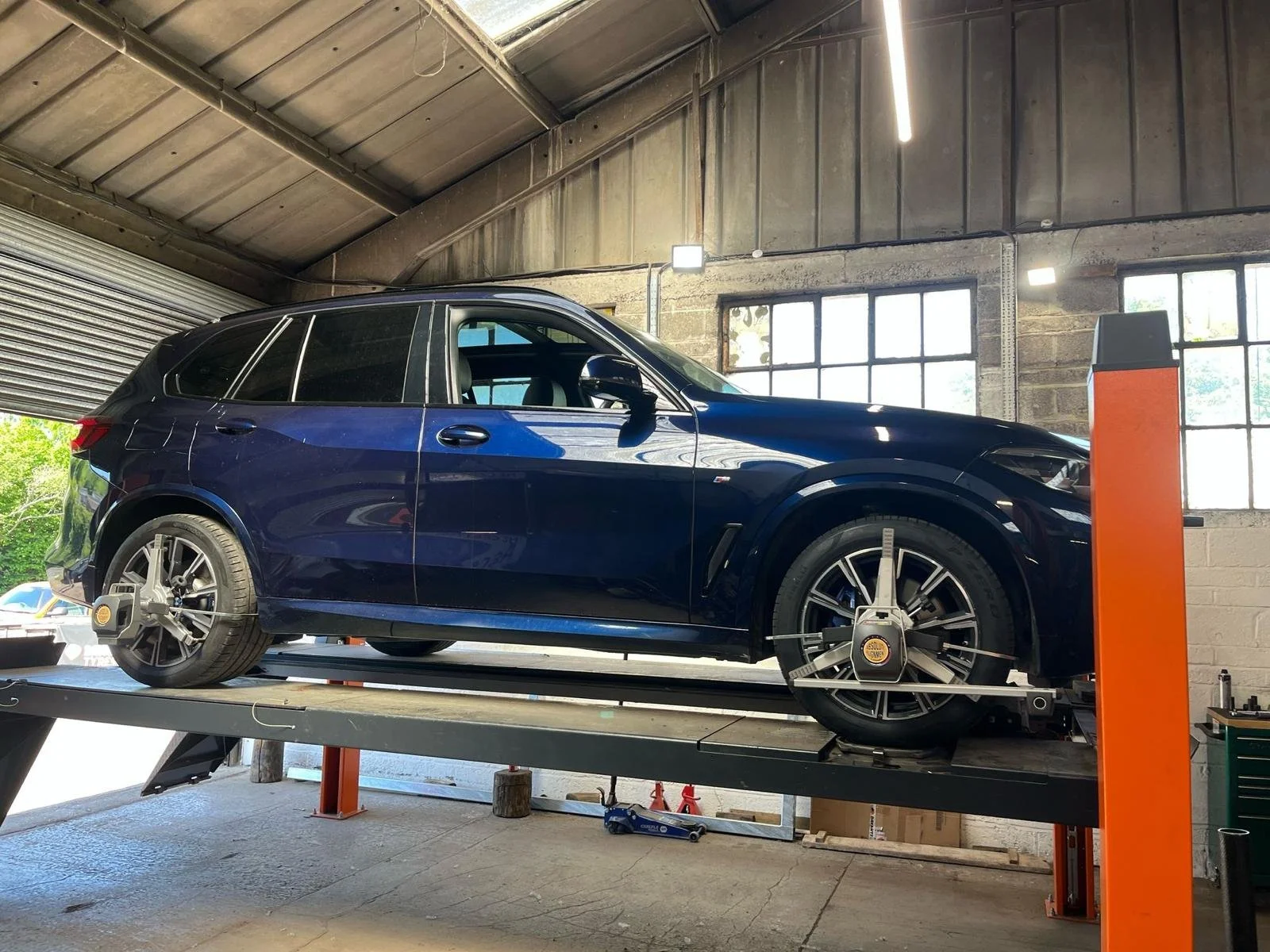 A blue SUV on a wheel alignment rack inside an industrial garage with large windows and exposed brick and metal walls.