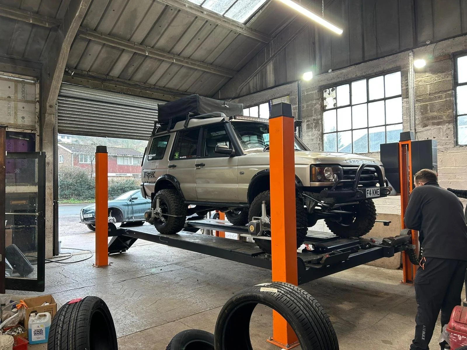 A dark-colored vehicle is positioned on a hydraulic lift inside an industrial-looking garage with large windows. A person is working near the vehicle, which appears to be undergoing maintenance or repair.