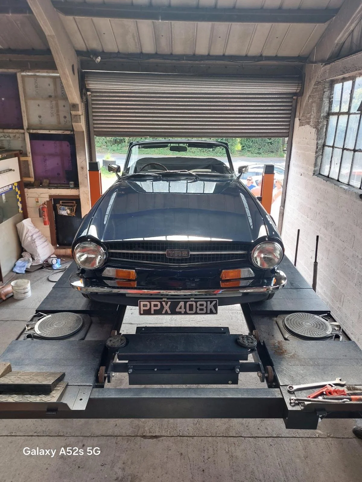 A black classic Triumph TR6 sports car with a license plate 'PPX 408K' is on a mechanical chassis dynamometer in a garage or workshop. The garage has a partially open roll-up door, concrete walls, and a window on the right side. There are various too