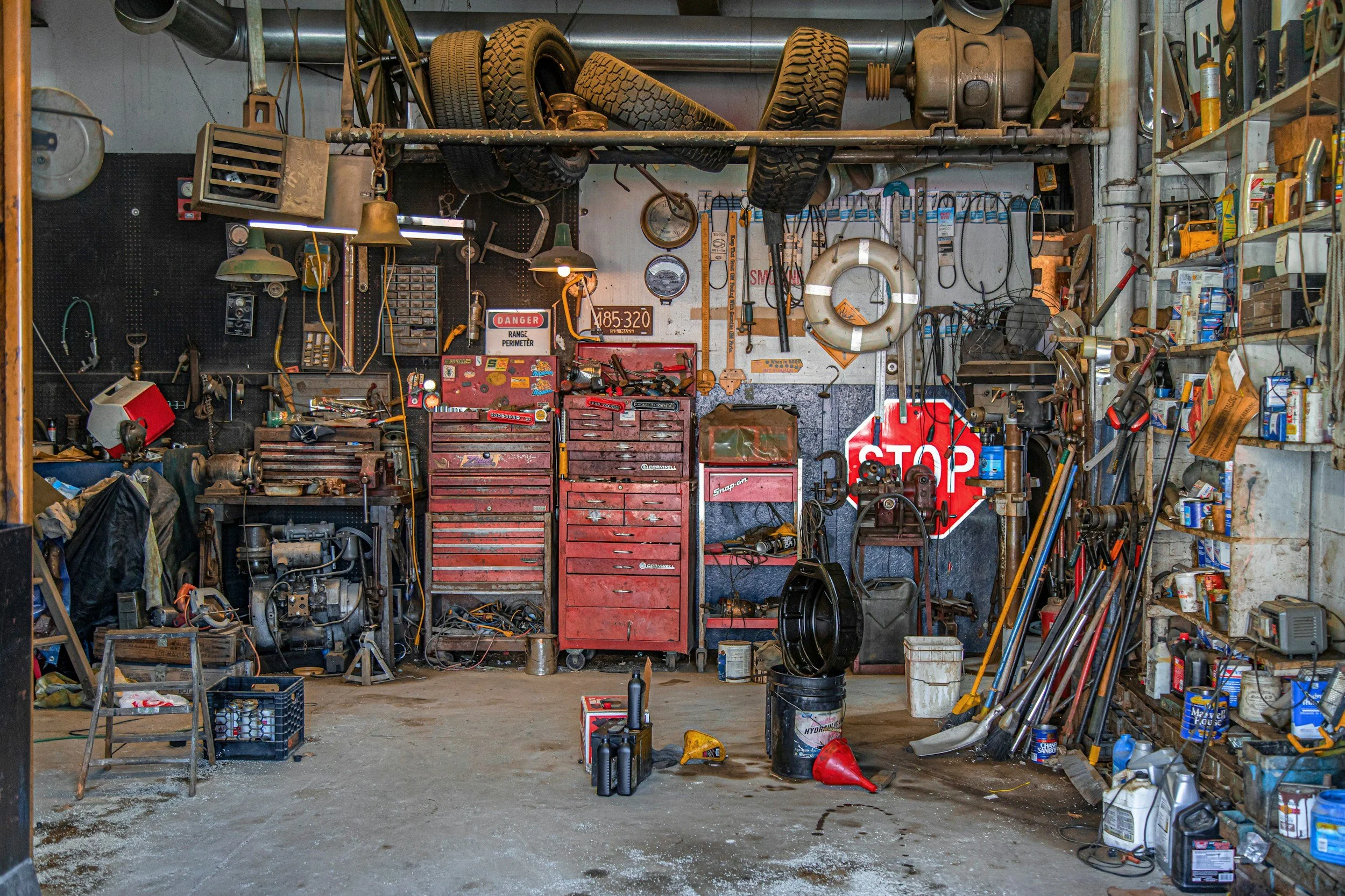 A cluttered garage with tires hanging from the ceiling, shelves filled with tools and supplies, and various items scattered on the floor and workbenches.