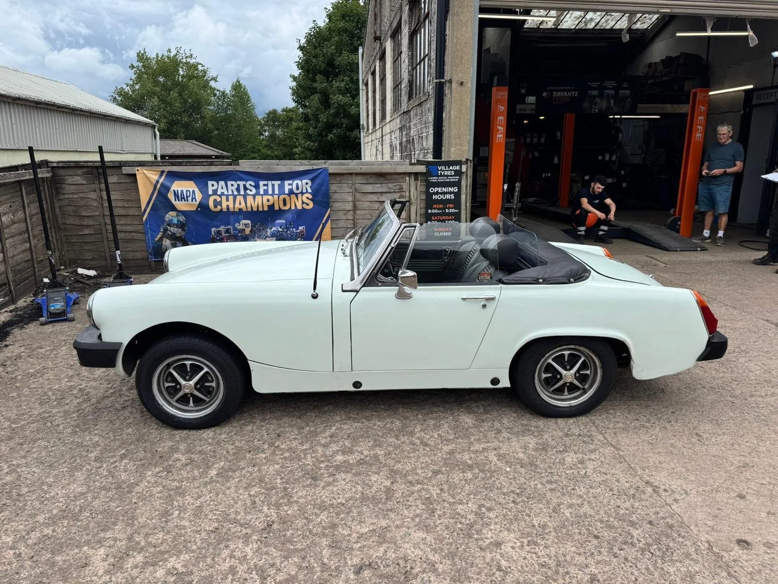 A white vintage convertible car parked outside an auto repair shop with a NAPA Parts sign in the background.