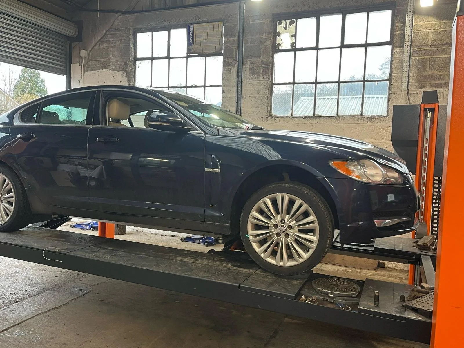 Black sedan car on a vehicle lift in a garage or auto repair shop with large windows and exposed brick wall.