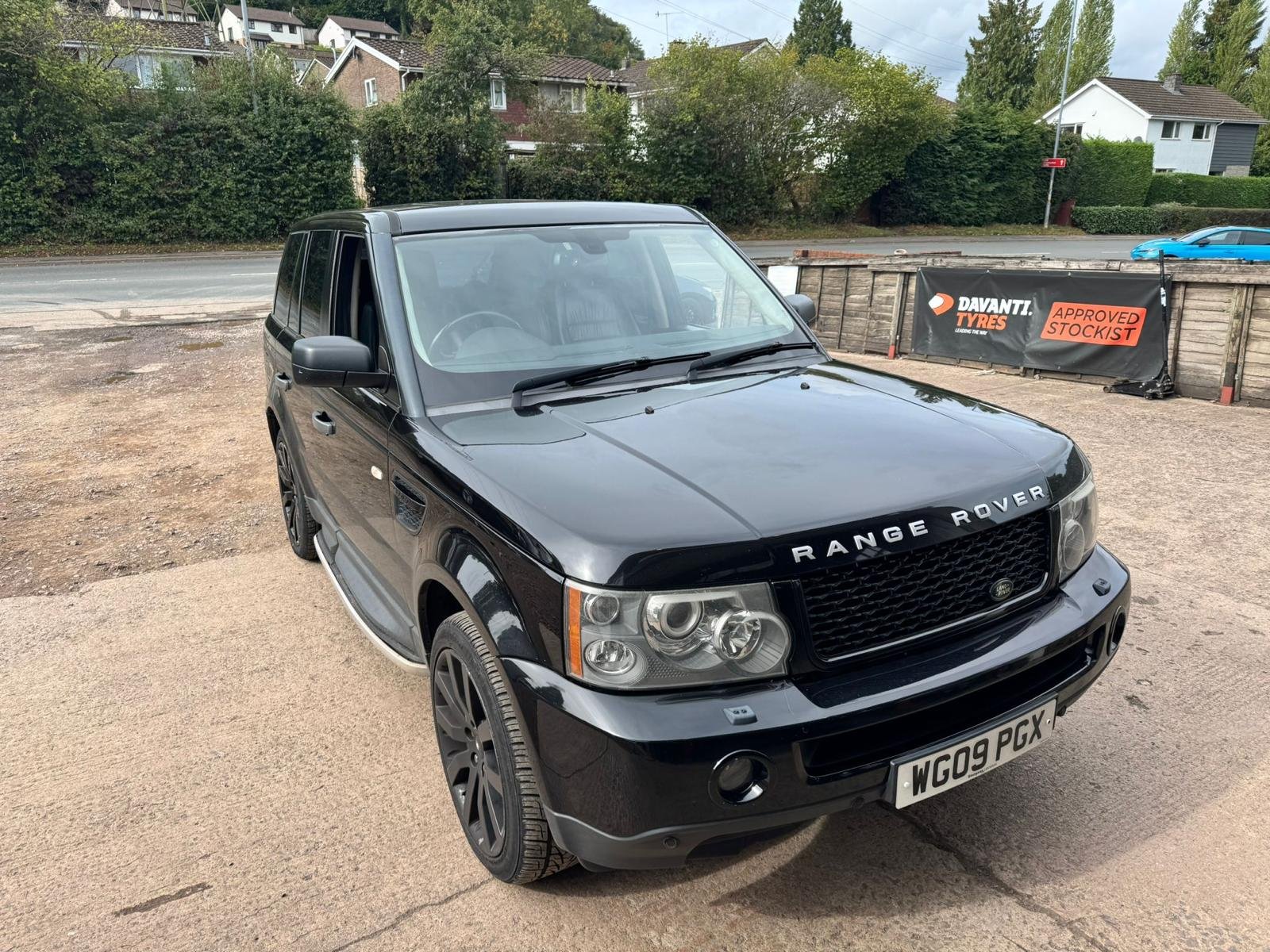 A black Range Rover parked on a gravel surface with residential houses and trees in the background. A sign advertising Davanti tires and approved stockist is visible behind the vehicle.