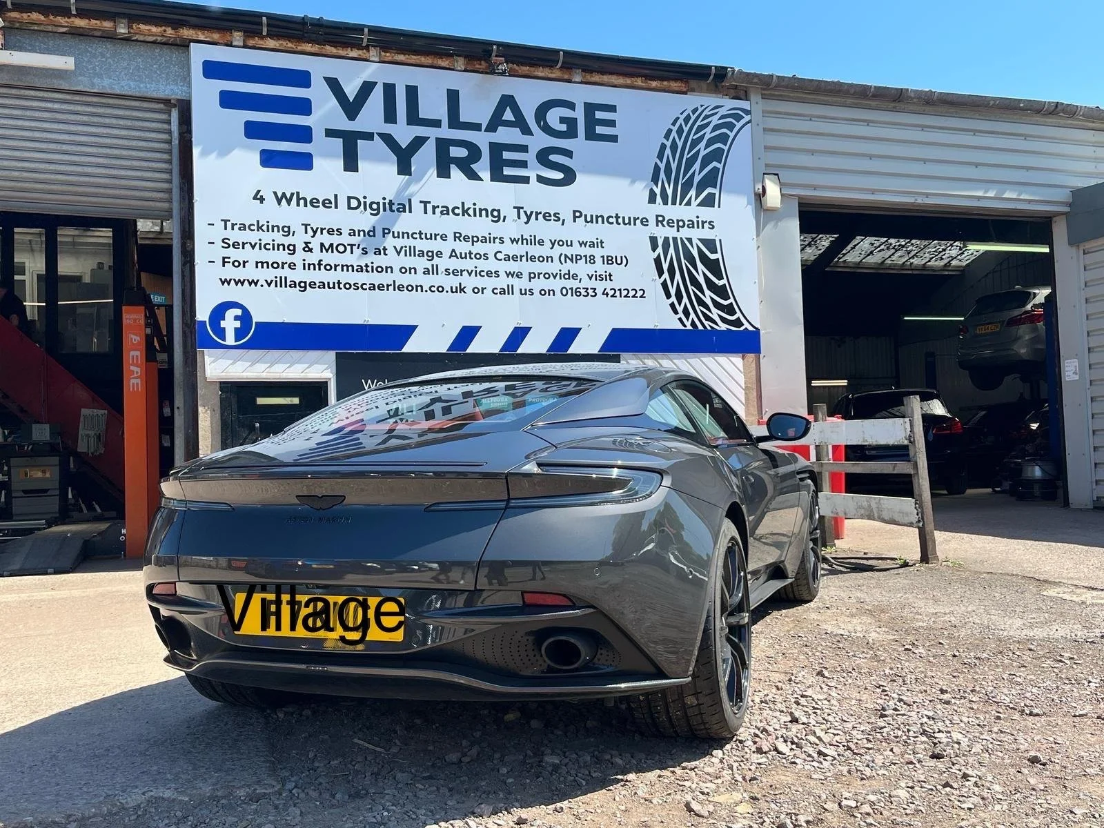A black Aston Martin car with a yellow license plate that reads 'Village' is parked in front of a tire shop. The shop has a large sign that says 'Village Tyres' and offers services including digital tracking, tyre repairs, puncture repairs, and MOTs.