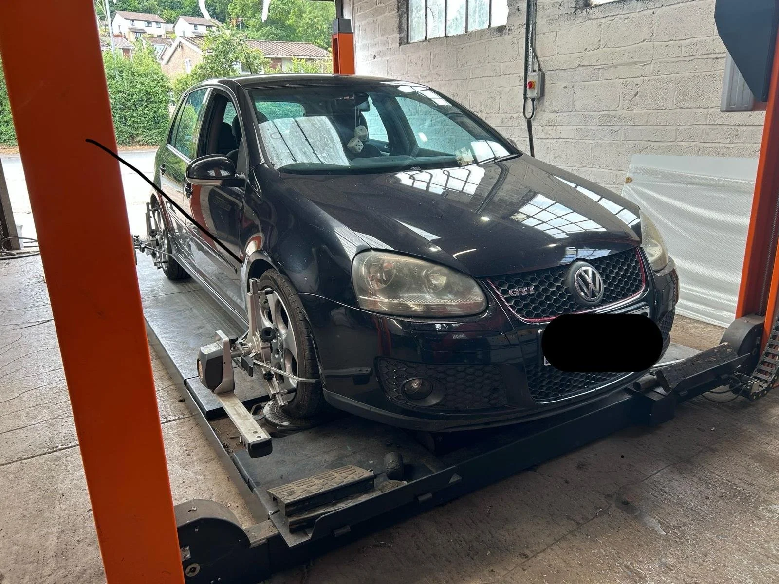 A black Volkswagen car on a wheel alignment machine inside a garage, with a brick wall and windows in the background.