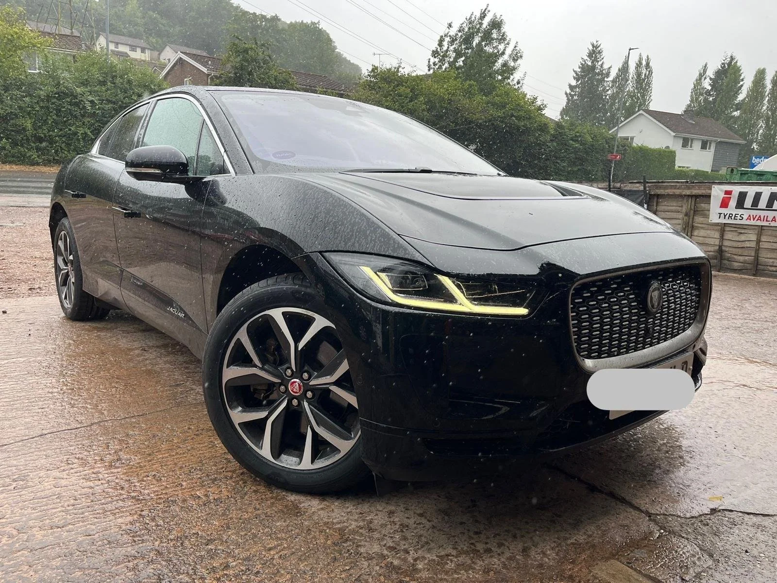 A black Jaguar electric vehicle parked on a muddy, wet surface during rainy weather, with residential houses and green trees in the background.