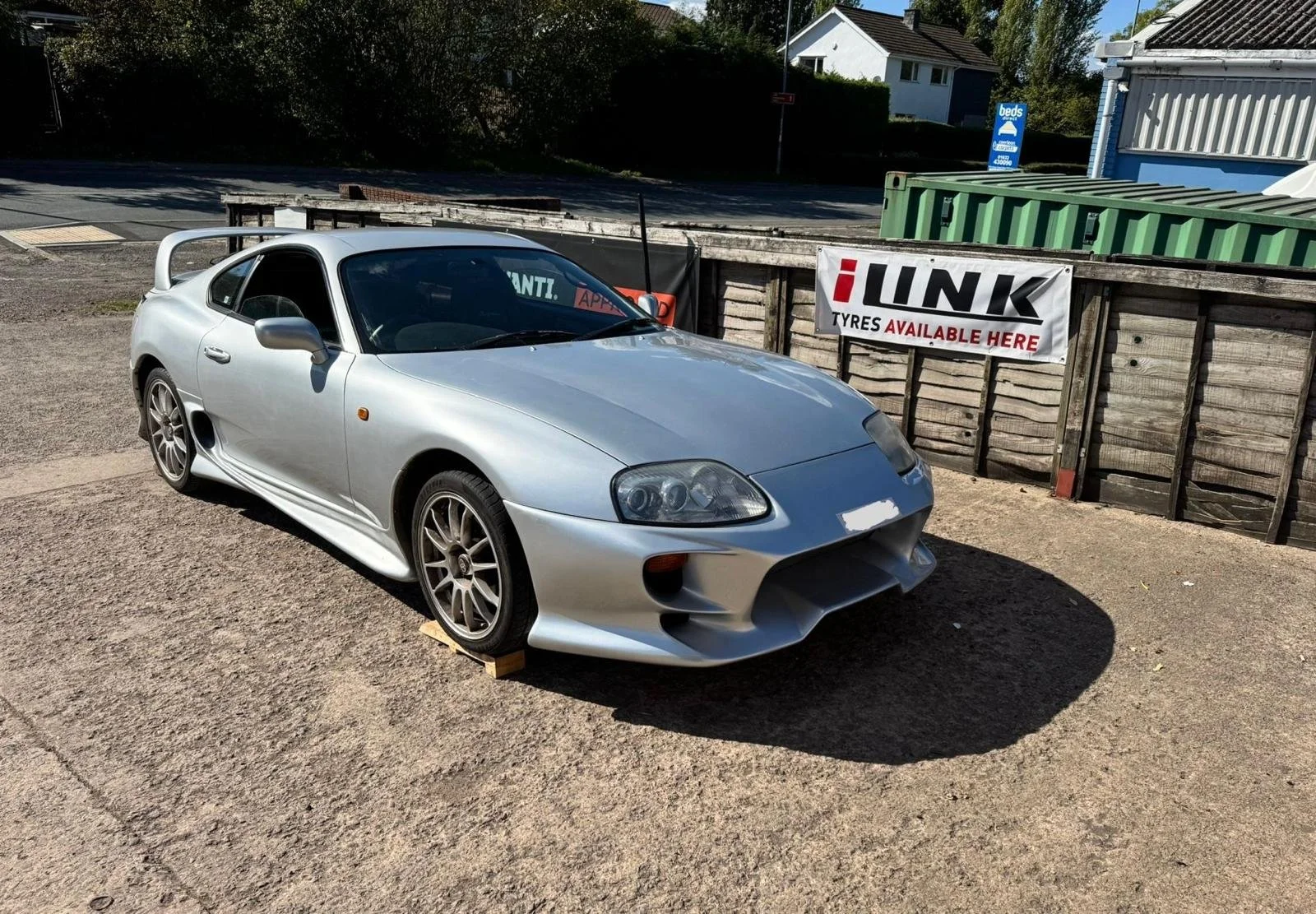 Silver Toyota Supra sports car parked on a gravel surface, supported by small wooden blocks under the front wheels, near a wooden fence with a sign advertising tire services, with houses and trees in the background.