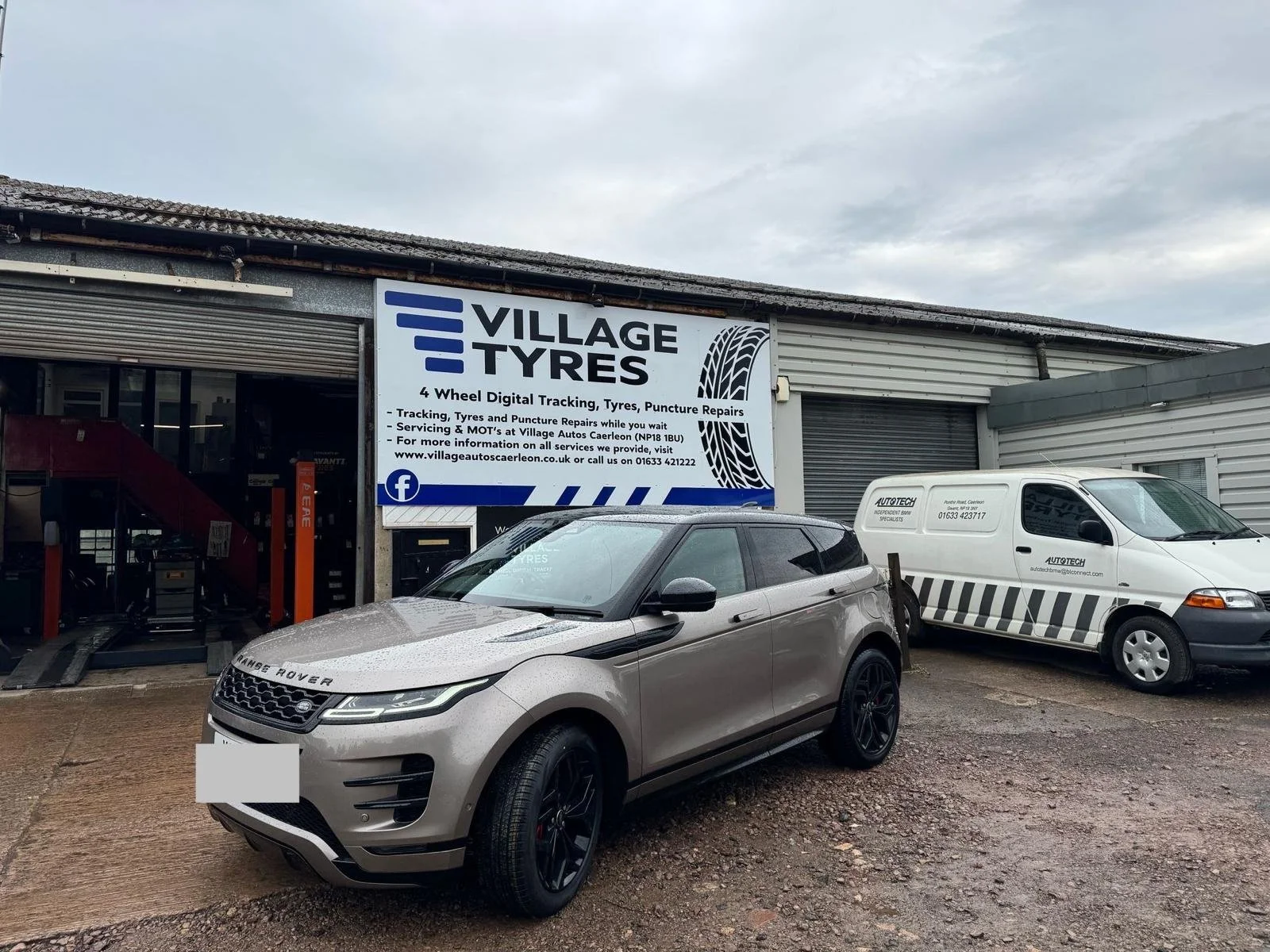 Exterior of Village Tyres with a large sign indicating tire and puncture repair services, two parked vehicles (a silver Range Rover and a white service van), and a partially open garage door.