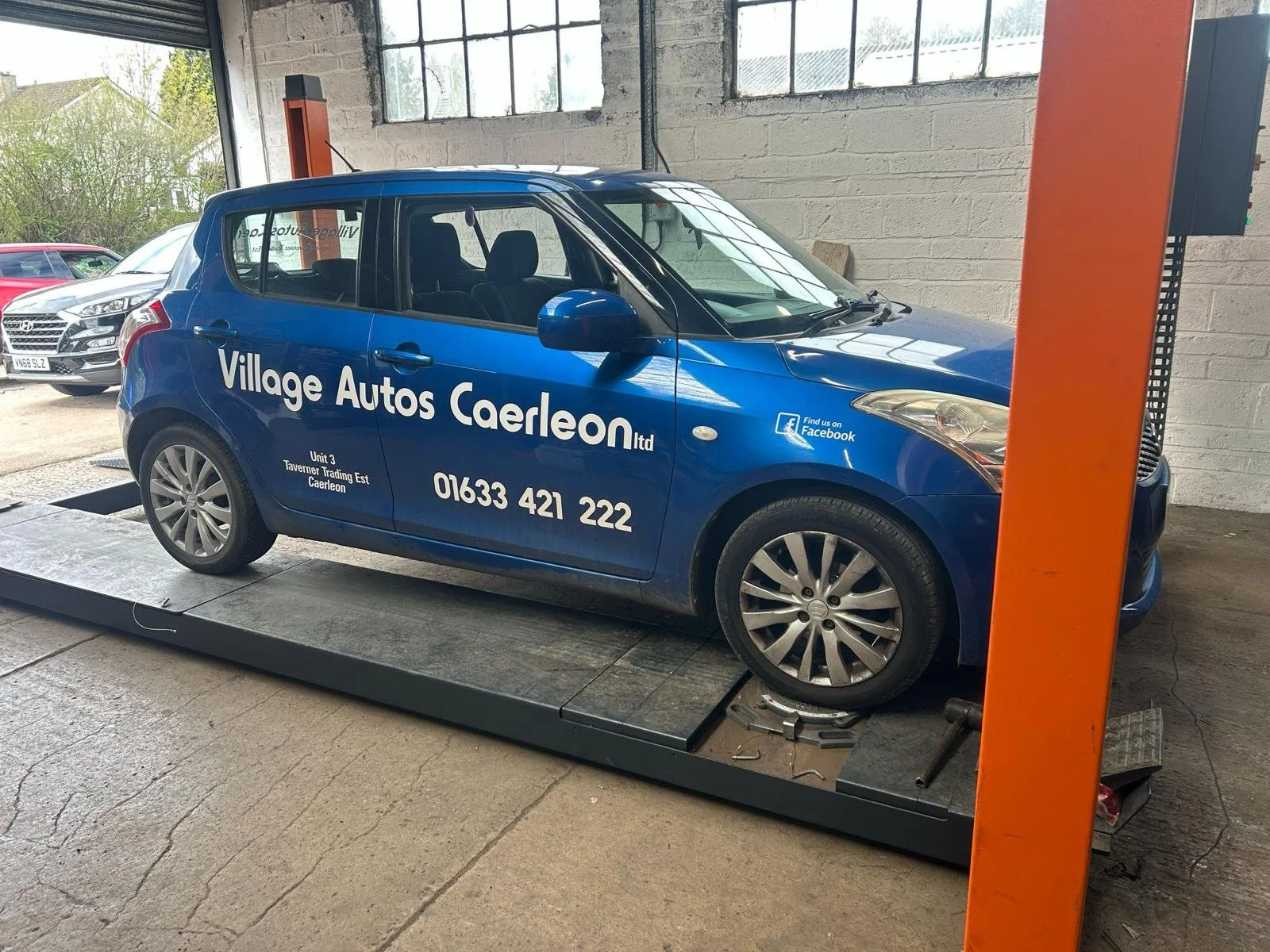 A blue car with advertising decals on its side, parked inside a garage or workshop, possibly on a lift or platform for maintenance or inspection.