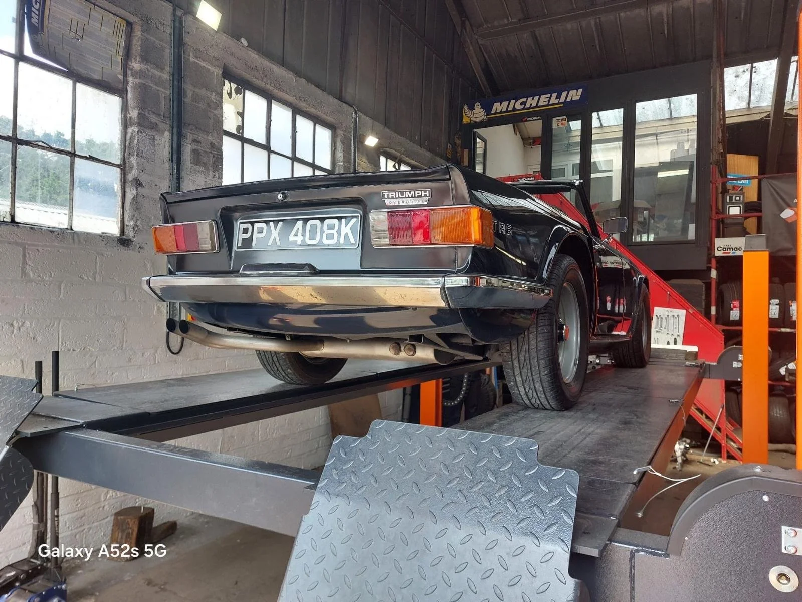 A vintage black Triumph sports car on a car lift in a garage. The car has a UK license plate 'PPX 408K' and is positioned for maintenance or display. The garage has large windows, metal walls, and various automotive signs and equipment.