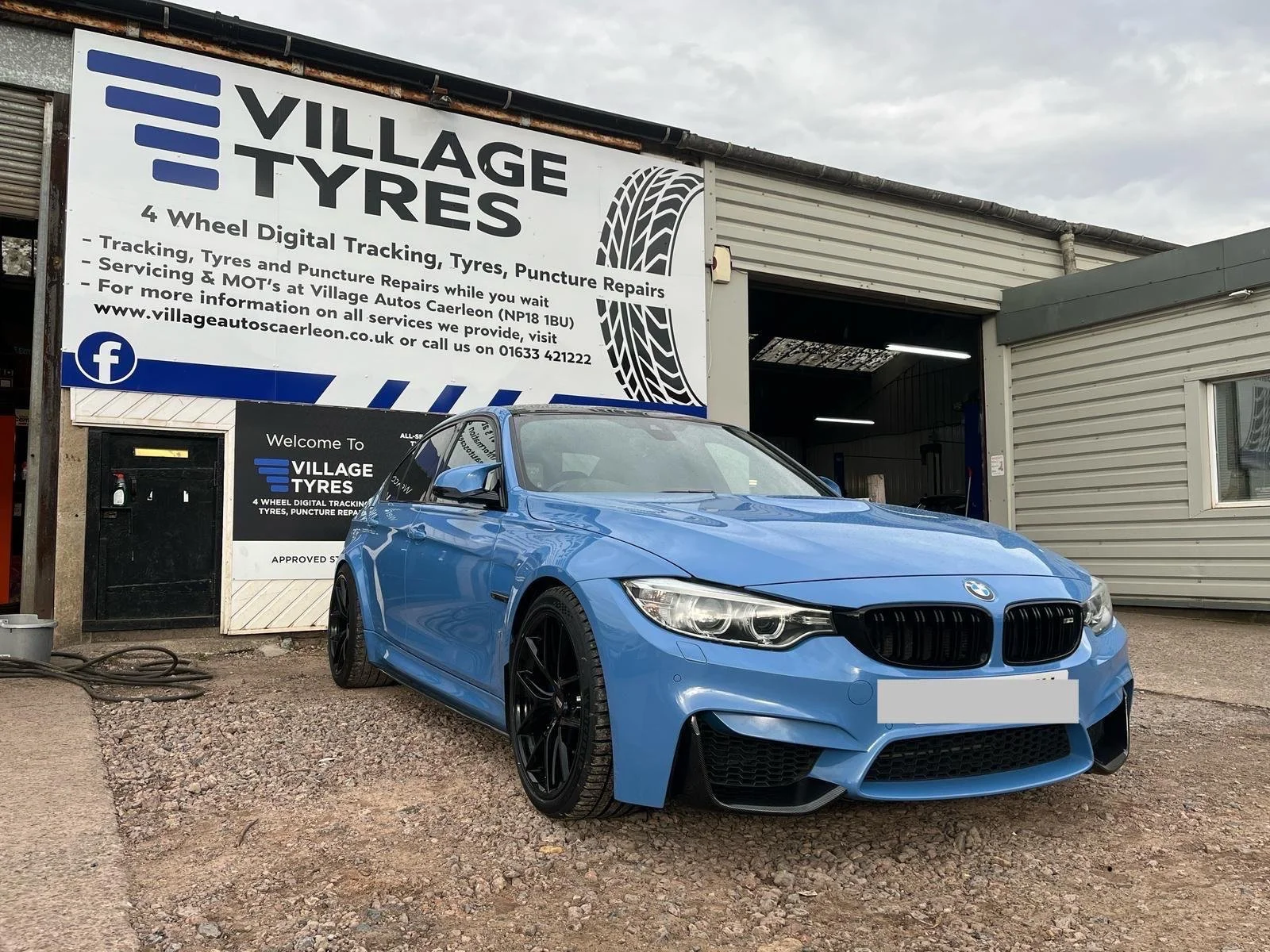 Blue BMW car parked outside Village Tyres auto repair shop with signage advertising tire tracking, puncture repairs, and MOT services.