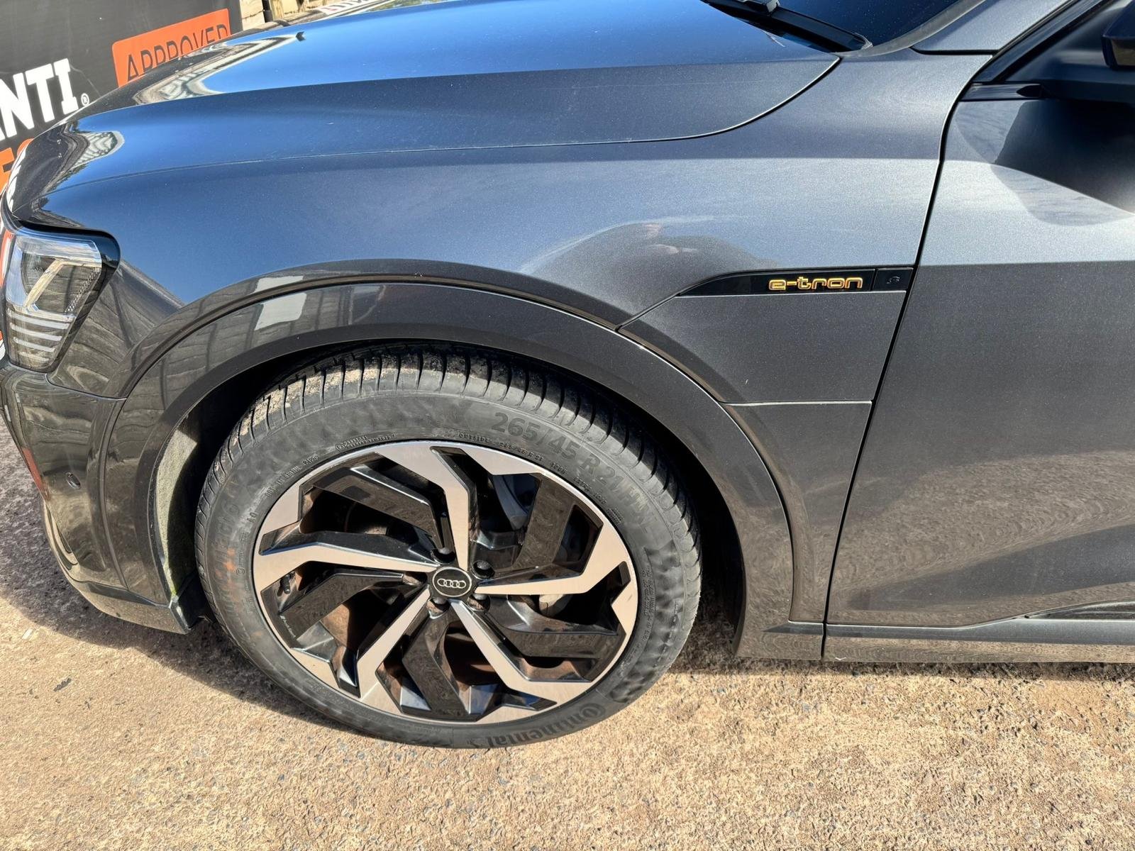Close-up of a dark gray Audi electric vehicle front left side, showing the wheel, tire, headlight, and e-tron badge.
