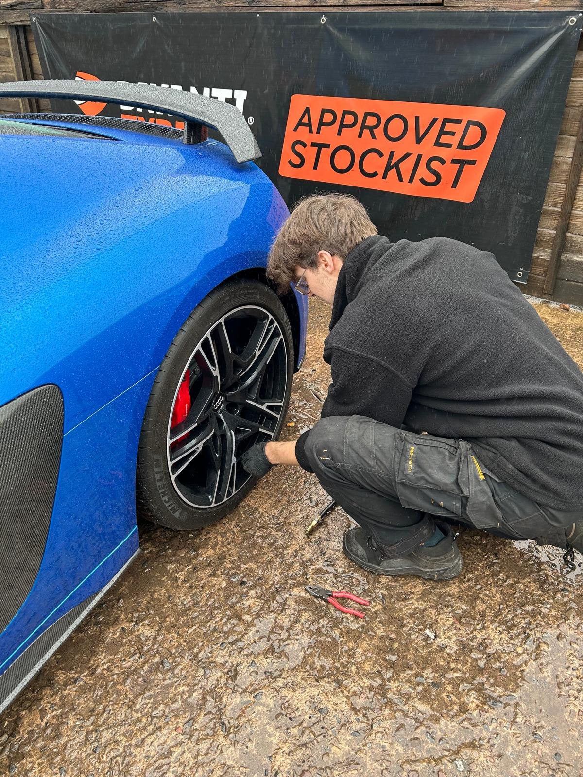 A man in black work clothes is kneeling next to a blue sports car, checking or repairing the front wheel, on a gravel surface. A sign behind him reads 'APPROVED STOCKIST'.