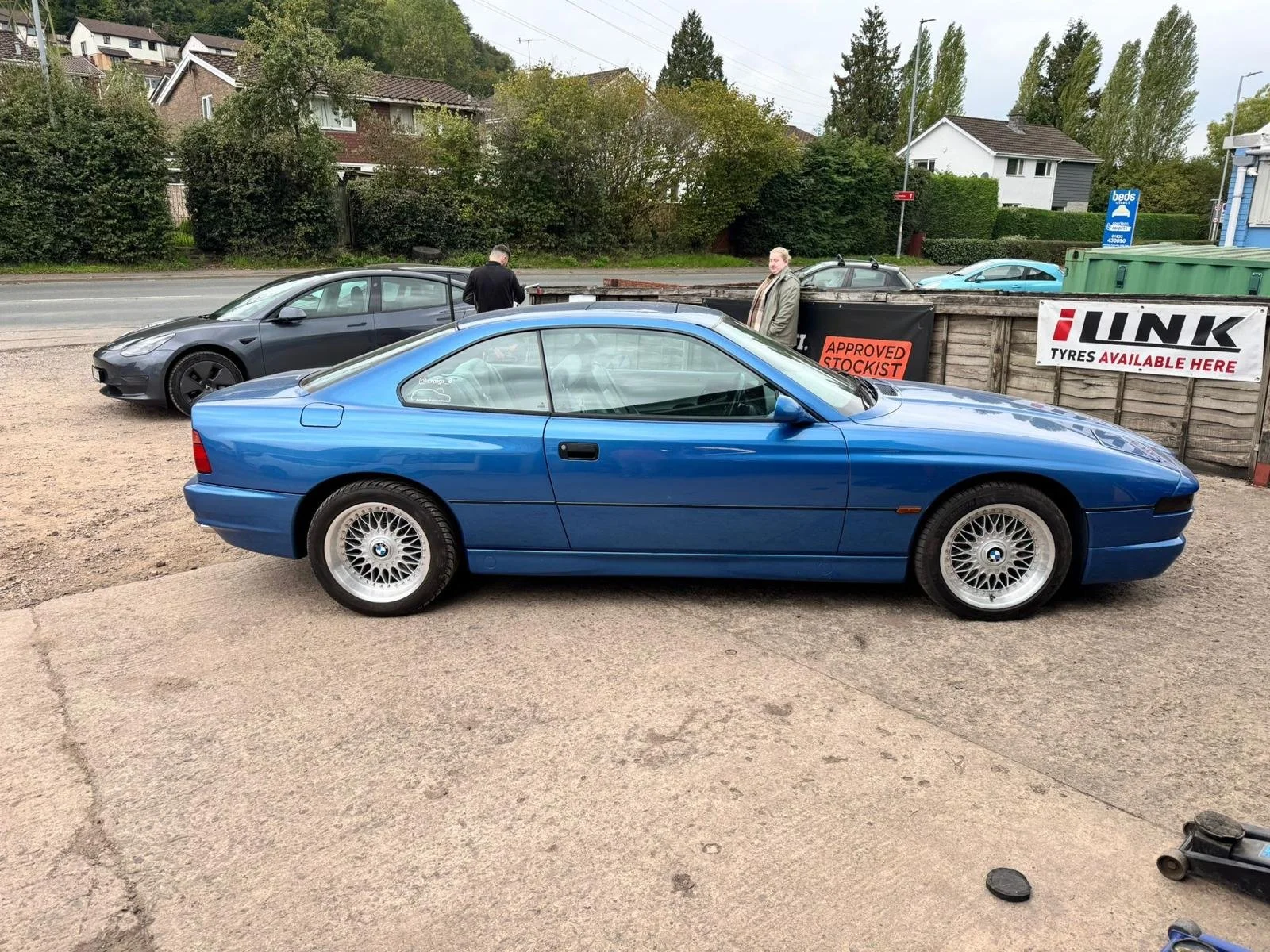 A blue BMW Z3 parked on a gravel lot with two men standing nearby, one looking at his phone and the other facing the camera. In the background, there are other cars, a fence with signs, and houses with trees.