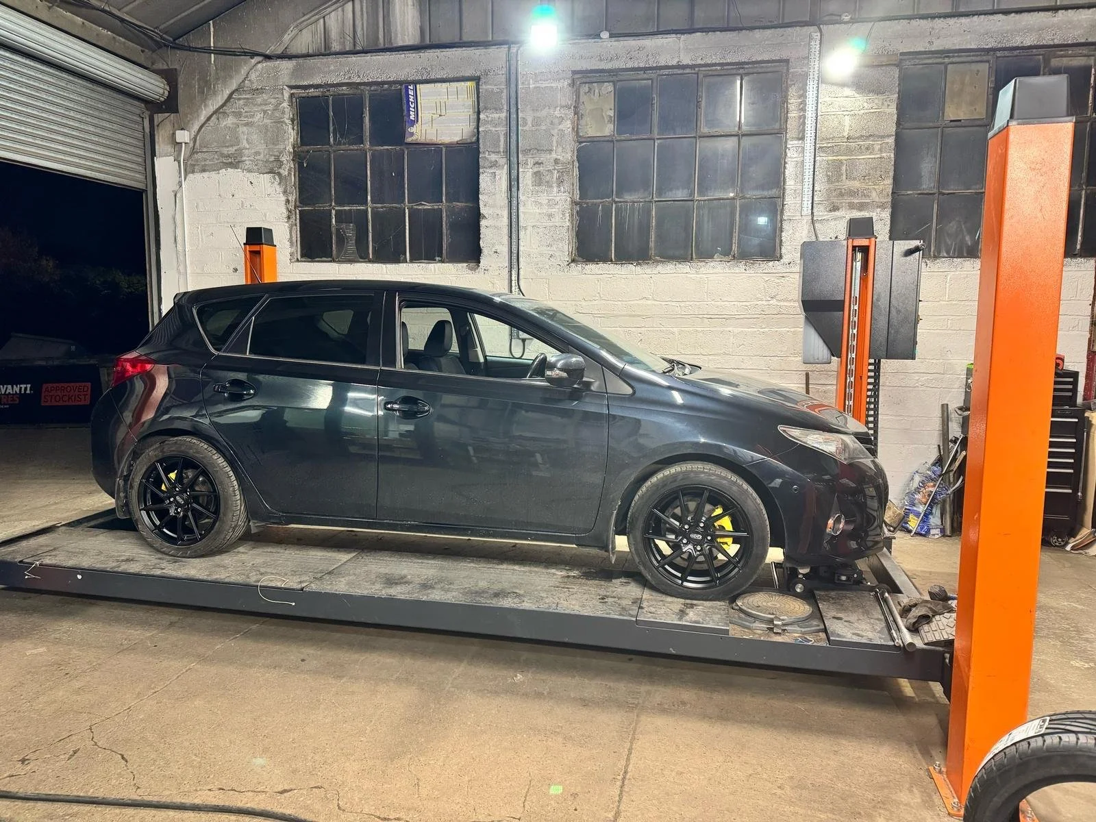 A black car is positioned on a vehicle dynamometer, with wheels on rollers, in an industrial garage with exposed brick walls and large windows. The garage is illuminated by a bright overhead light.