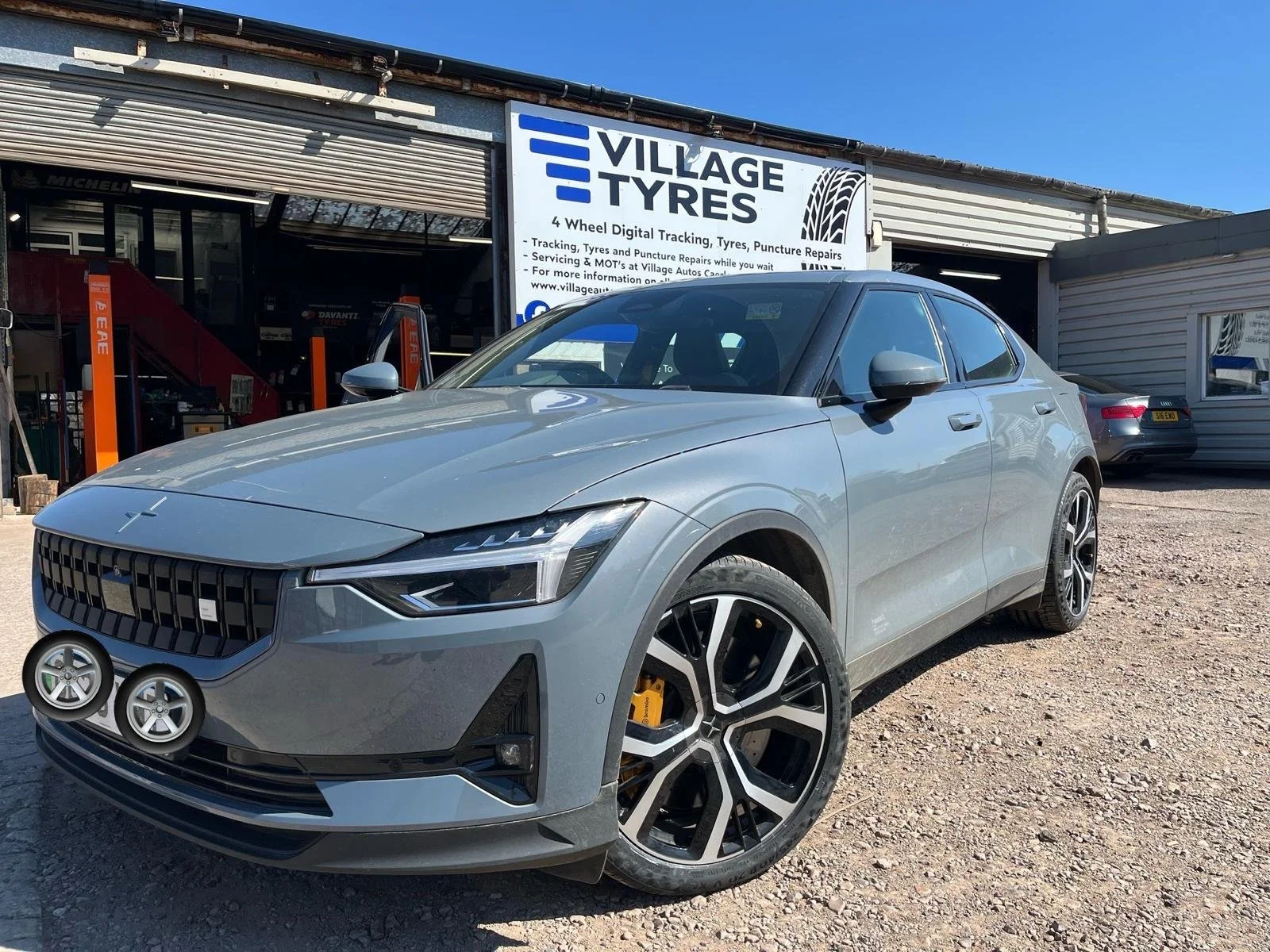 A silver modern hatchback car parked outside of a Tyre shop with a sign that says 'Village Tyres', featuring a black and white tire graphic, on a sunny day with a clear blue sky.